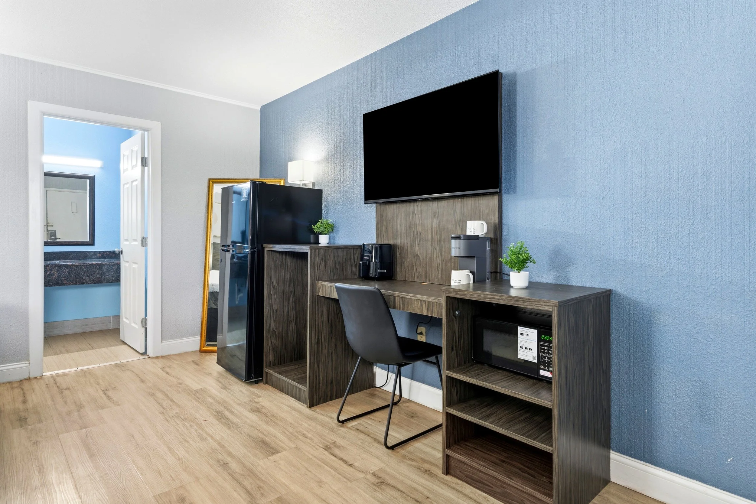 A hotel or apartment room with a wall mounted flat-screen TV, a small desk with a black chair, a mini refrigerator, microwave, coffee maker, and two small potted plants. An open door shows a bathroom with a blue wall and a mirror.