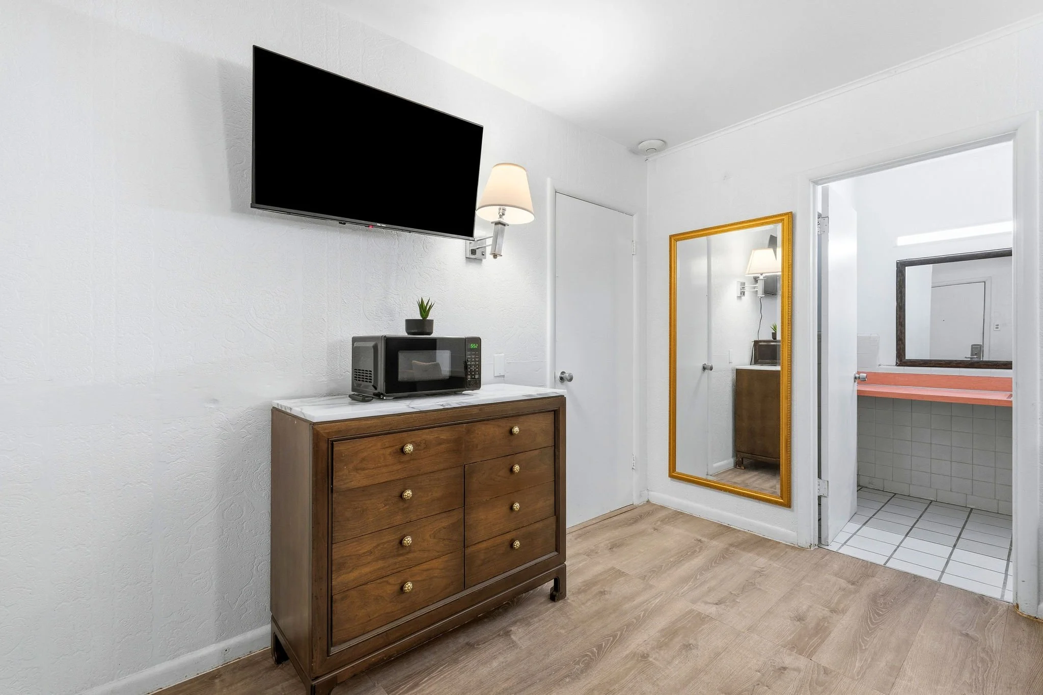 Inside a hotel room, a flat-screen television mounted on a white wall, a small microphone on a wooden dresser with a potted plant, a microwave oven, and a large gold-framed mirror reflecting a doorway and other parts of the room.