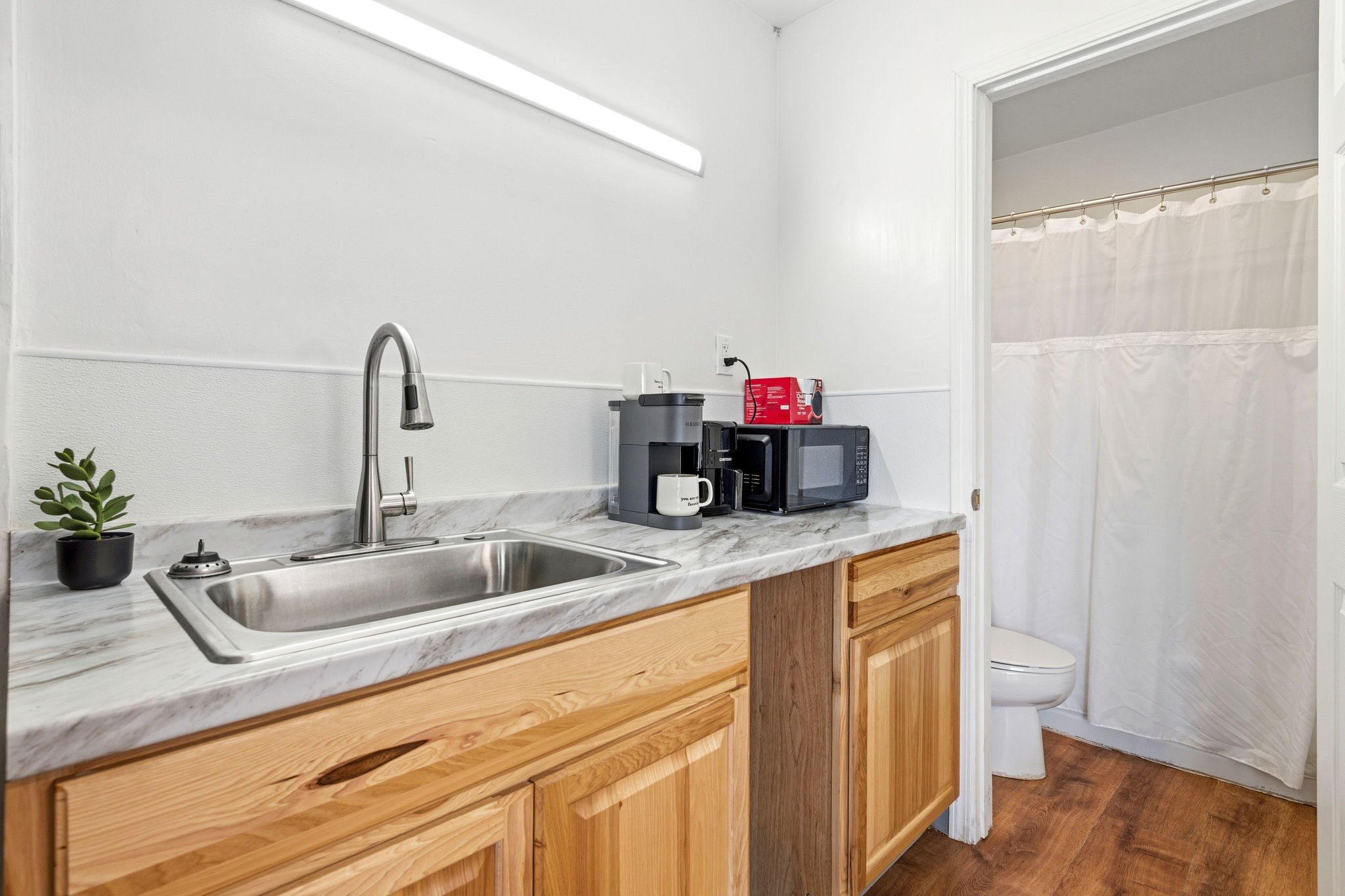 A kitchen with a marble countertop, wooden cabinets, a sink with a faucet, a small plant in a pot, a coffee maker, microwave, and a toaster. There is a doorway with a white curtain leading to a bathroom with a toilet.