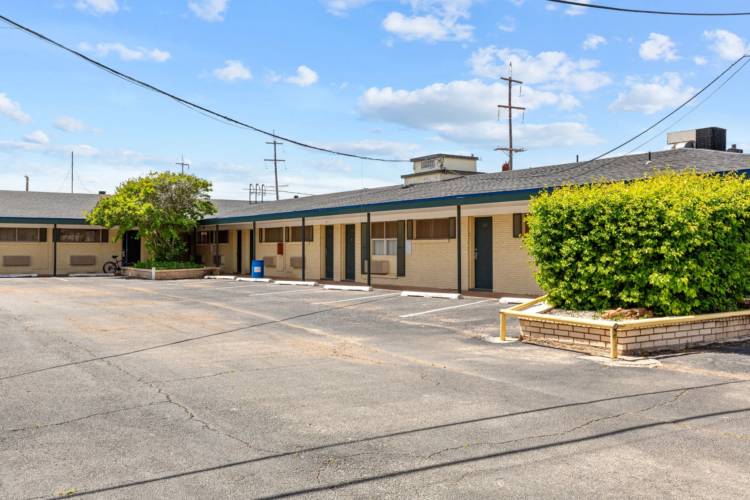 A single-story motel with yellow brick walls and a flat roof, with a parking lot in front. There are small trees and bushes around the building, and a bicycle is parked near one of the plants. Utility poles and power lines are visible against a blue 