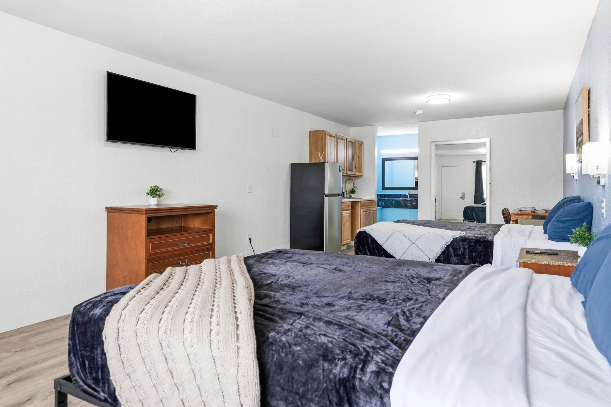 Interior of a room with two beds with dark blue velvet bedding and white pillows, a wooden dresser with a small potted plant, a kitchen area with wooden cabinets, a refrigerator, and a sink, and a living area with a white wall, a wall-mounted TV, and