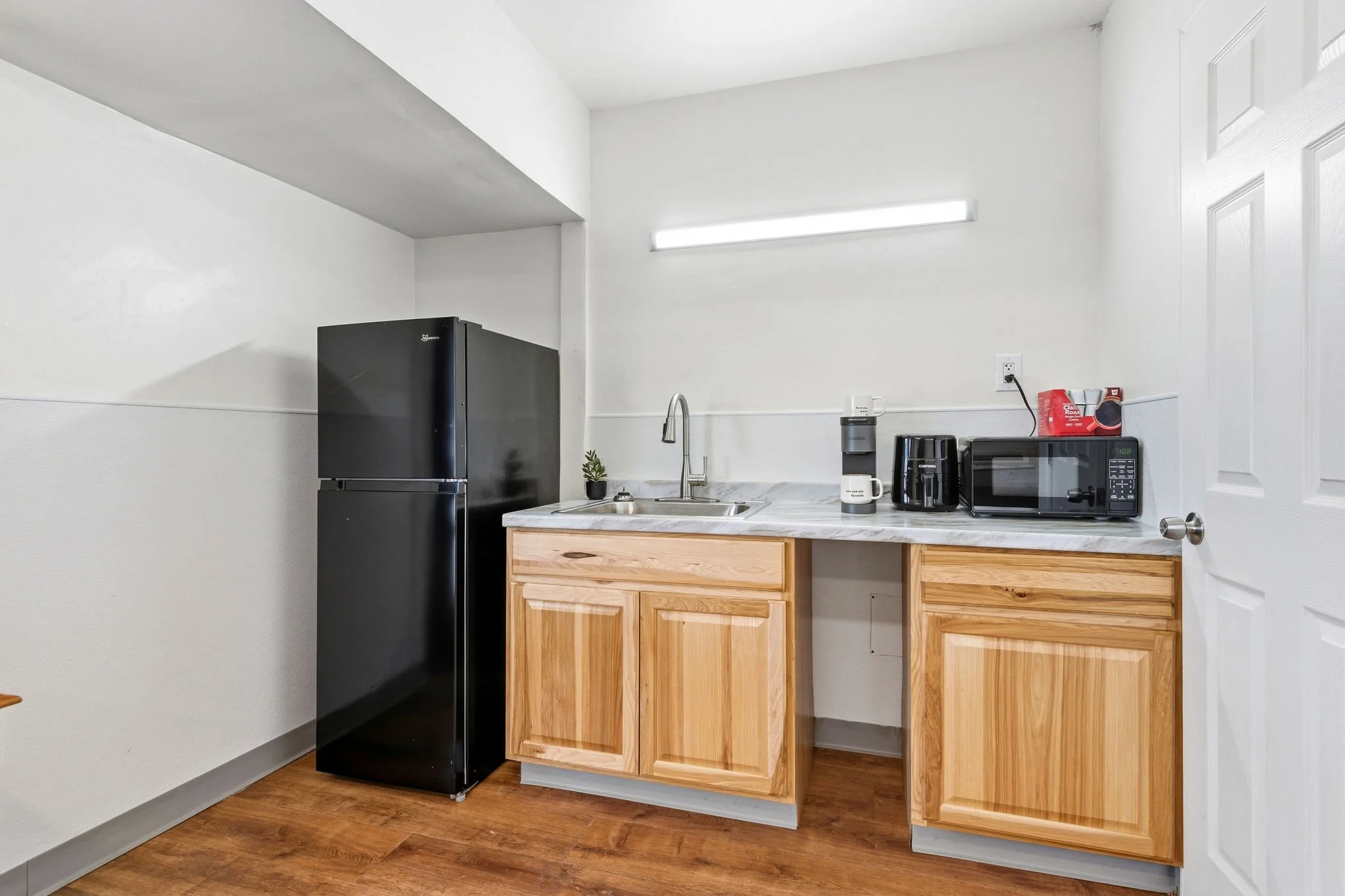Small kitchenette with wooden cabinets, countertop, sink, black refrigerator, microwave, coffee maker, water dispenser, white walls, and hardwood floor.