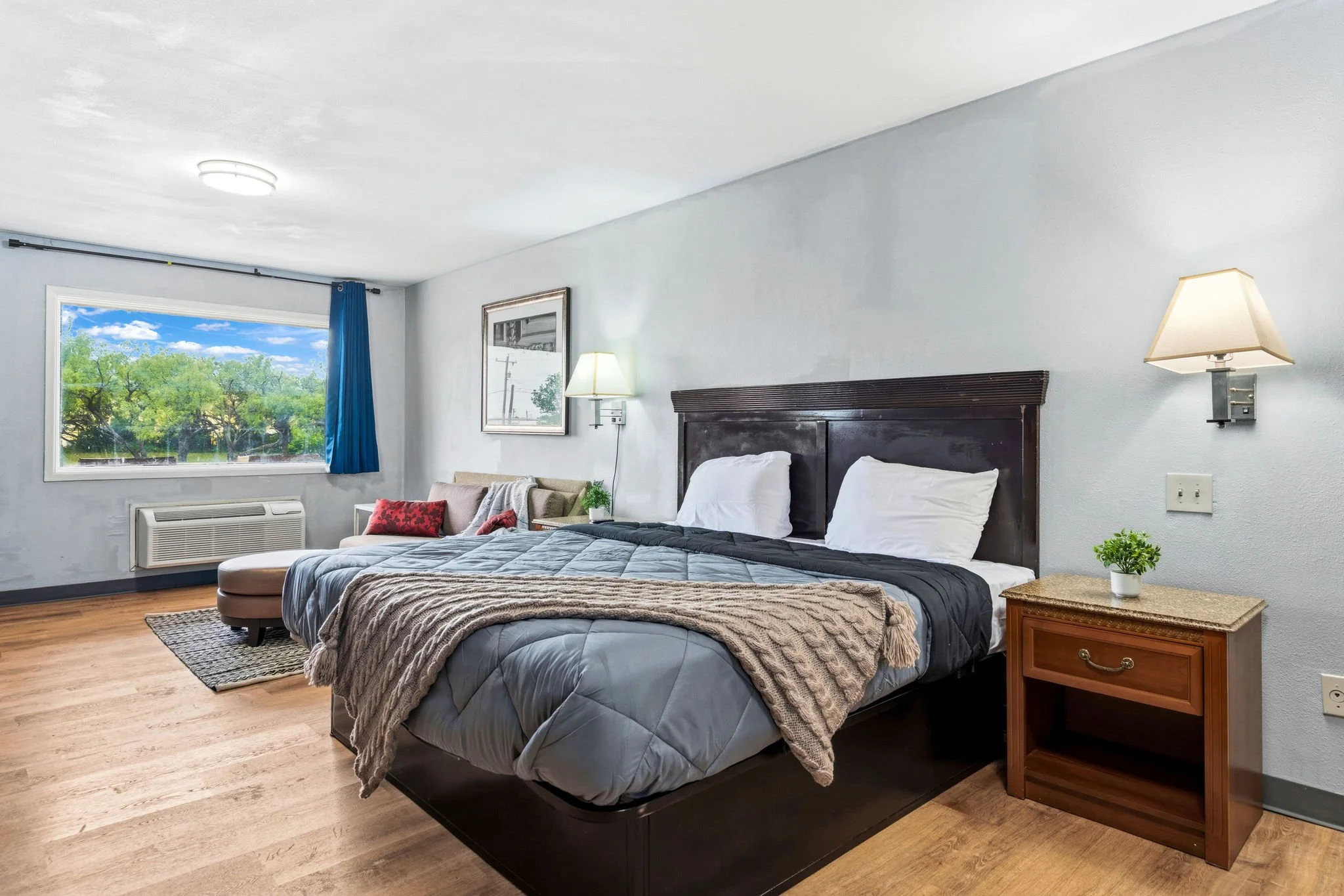 A hotel room with a large window showing green trees outside, a bed with dark wooden headboard, white pillows, gray and beige bedding, a beige armchair with red pillows, two wall-mounted lamps, a small bedside table with a potted plant, and light-col