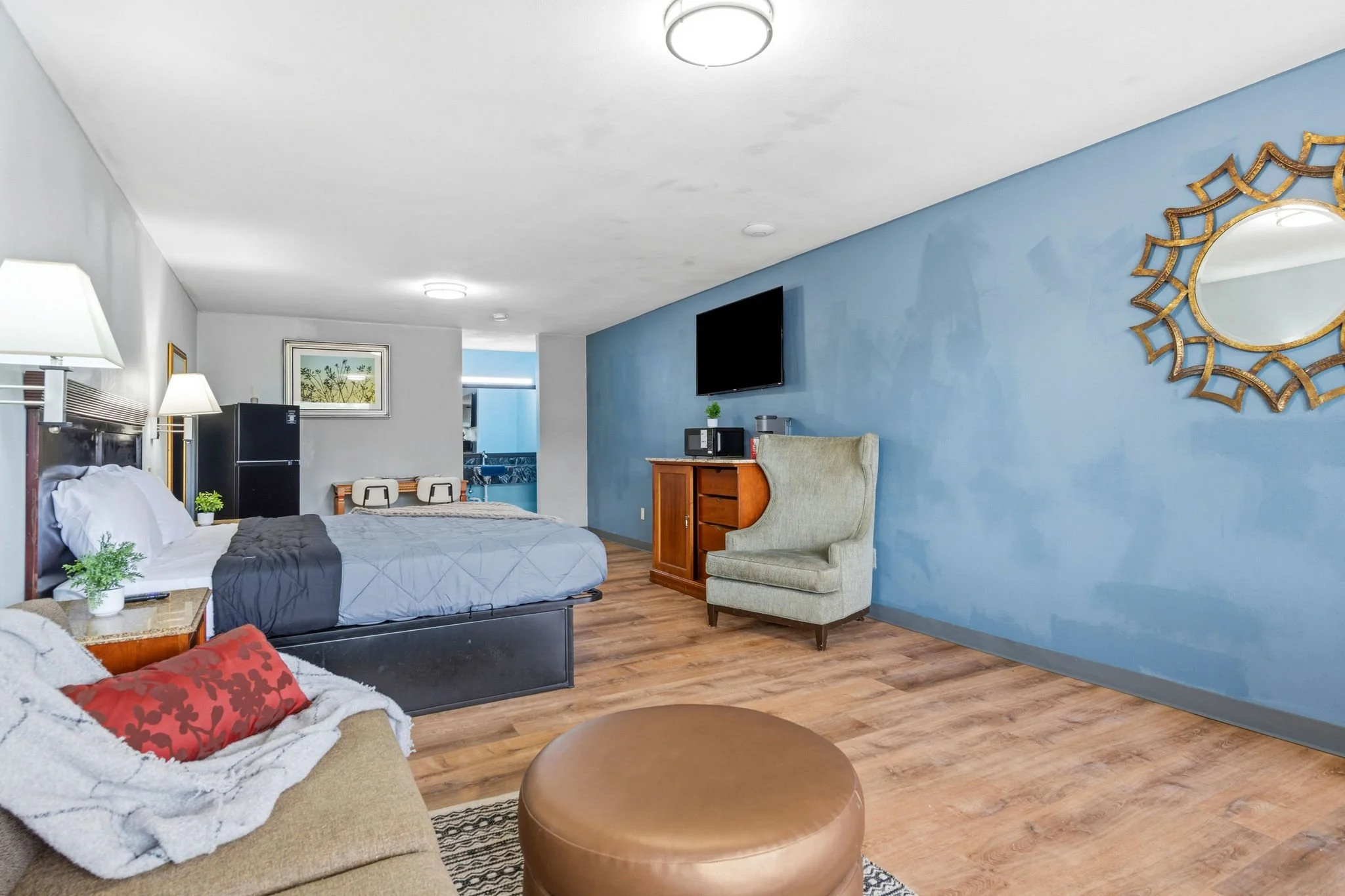 A hotel room with a large bed, a beige armchair, a small refrigerator, a microwave, a flat-screen TV, and decorative mirrors on the blue wall. There are lamps and framed pictures, with a wooden floor.