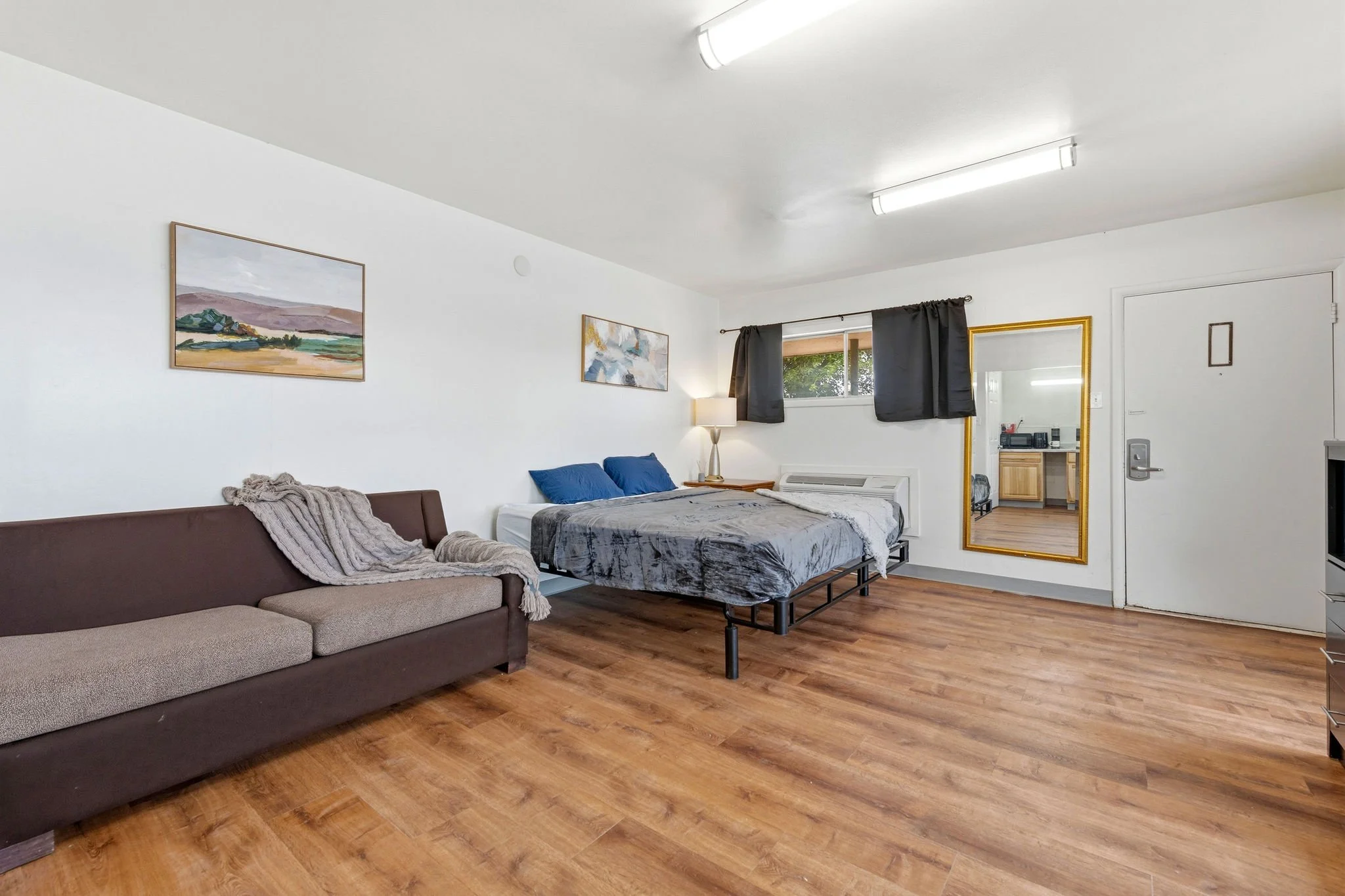 A studio apartment with white walls, hardwood floors, a brown sofa with beige cushions, a bed with gray and blue bedding, wall art, a window with black curtains, a mirror with a gold frame, a lamp, and a doorway leading to another room.
