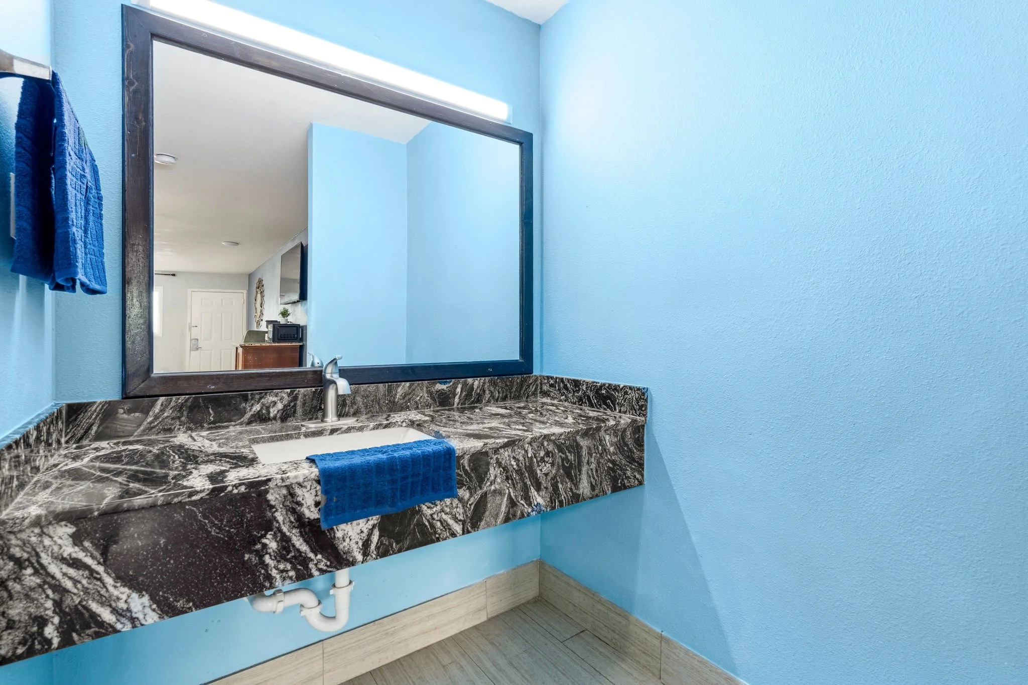 A bathroom vanity with a marble countertop, silver faucet, and an oval sink. A blue towel hangs on a rack above the counter. A large mirror is mounted on a light blue wall, reflecting part of a room with a door, a TV, and a small table with a decorat