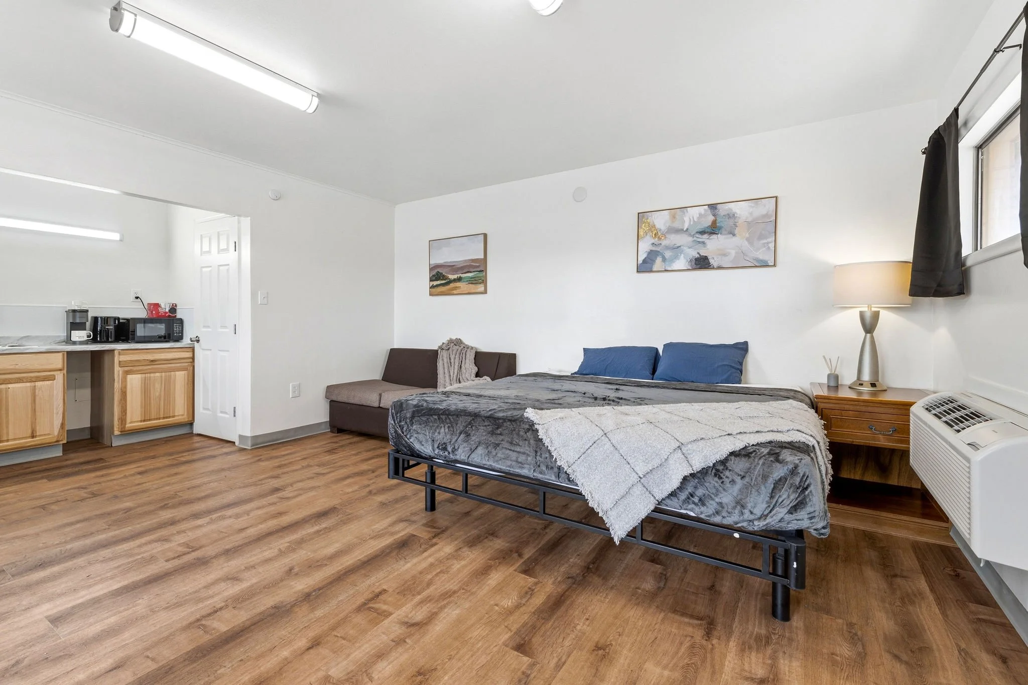 A bedroom with a bed, lamp, artwork on the wall, and a small living room area with a couch, kitchenette with microwave, coffee maker, and refrigerator, and wooden flooring.