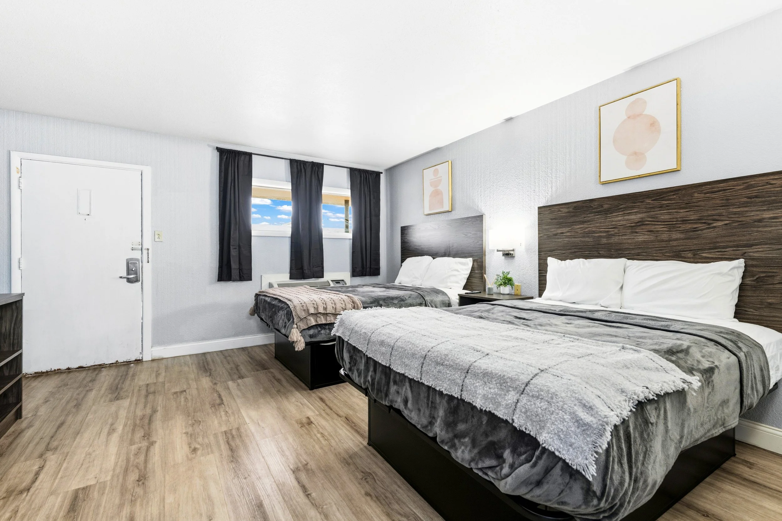 Two beds in a hotel room with dark wood headboards, white pillow and bedding, covered with blankets. There are two windows with black curtains, a small nightstand with a plant, and abstract artwork on the wall. Light wood flooring and white walls.