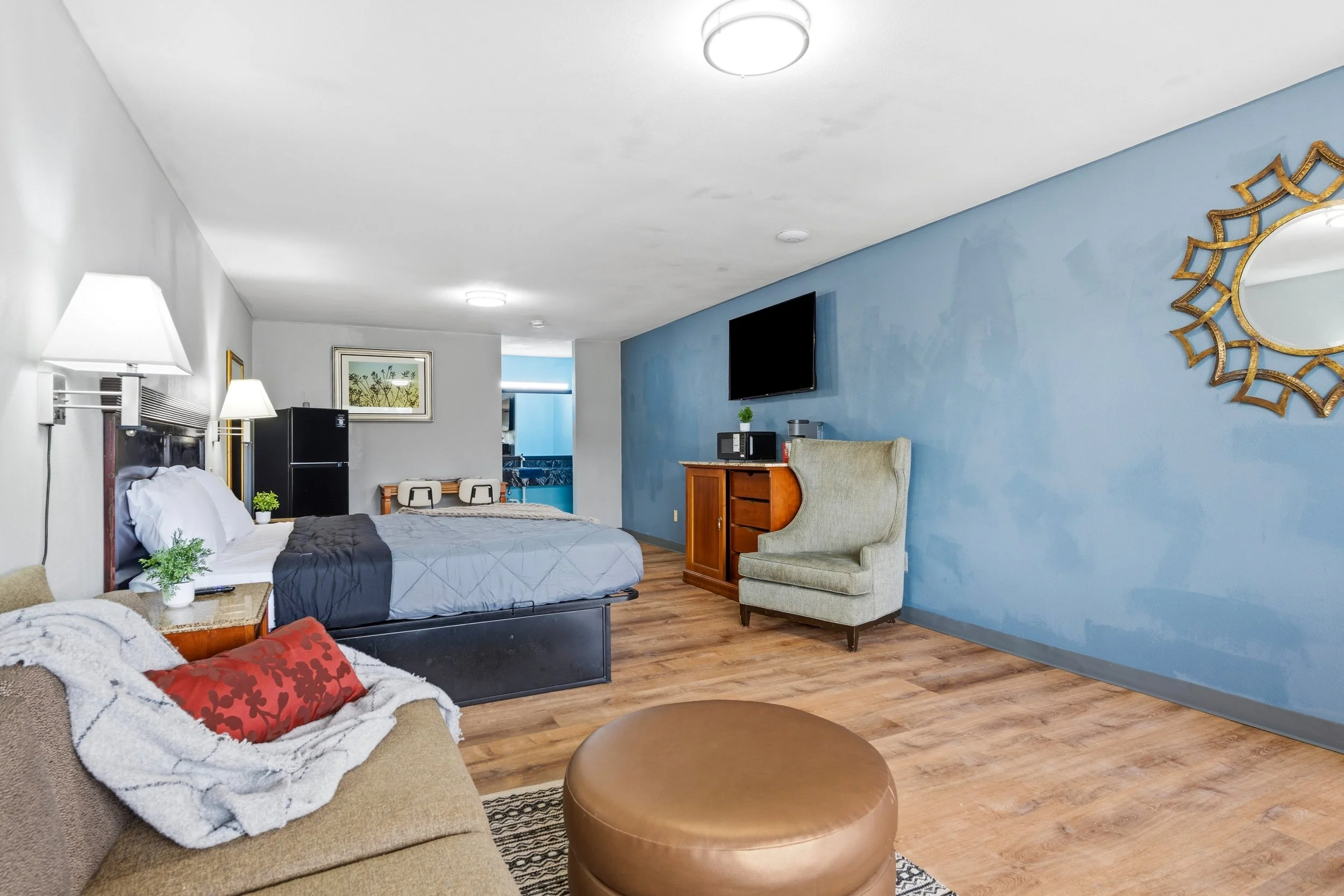 Inside a hotel room with a bed, a beige sectional sofa with red and white pillows, a small round coffee table, and a blue accent wall with a decorative round mirror. There's a flat-screen TV, a microwave, a small refrigerator, and a plant on a wooden cabinet. The room has wooden flooring and ceiling lights.