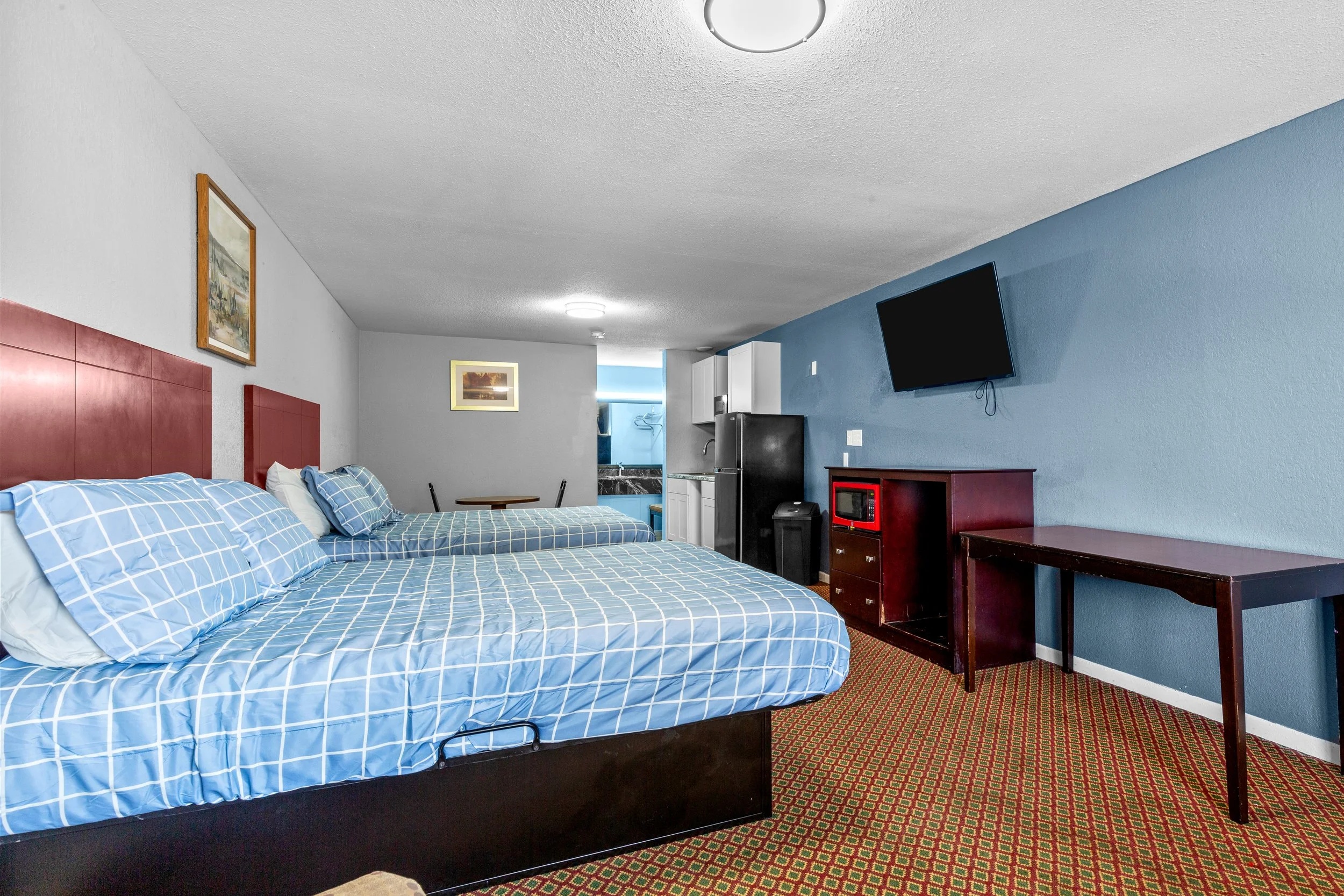 Hotel room with two beds covered in blue checkered bedding, a red headboard, framed artwork on the white wall, a small wooden table, a flat-screen TV on the blue accent wall, a dresser, a microwave, refrigerator, and kitchen area in the background, and patterned carpet.