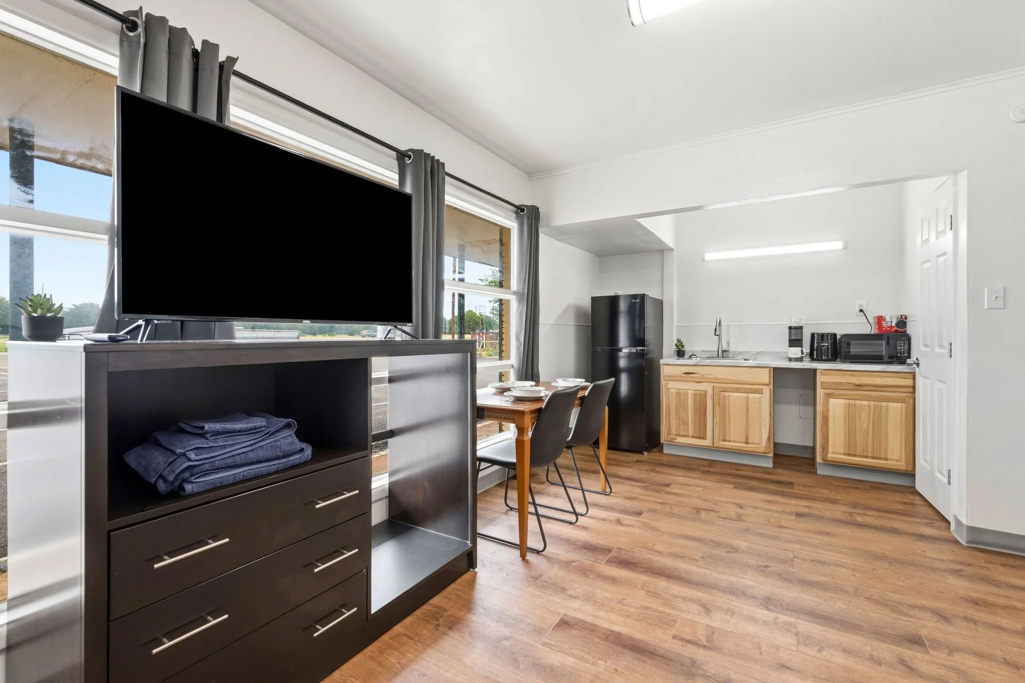 Interior of a modern apartment with a living area and kitchen, featuring wooden flooring, a black TV, a small black cabinet with folded towels, a dining table with chairs, a black refrigerator, and wooden kitchen cabinets with various appliances.