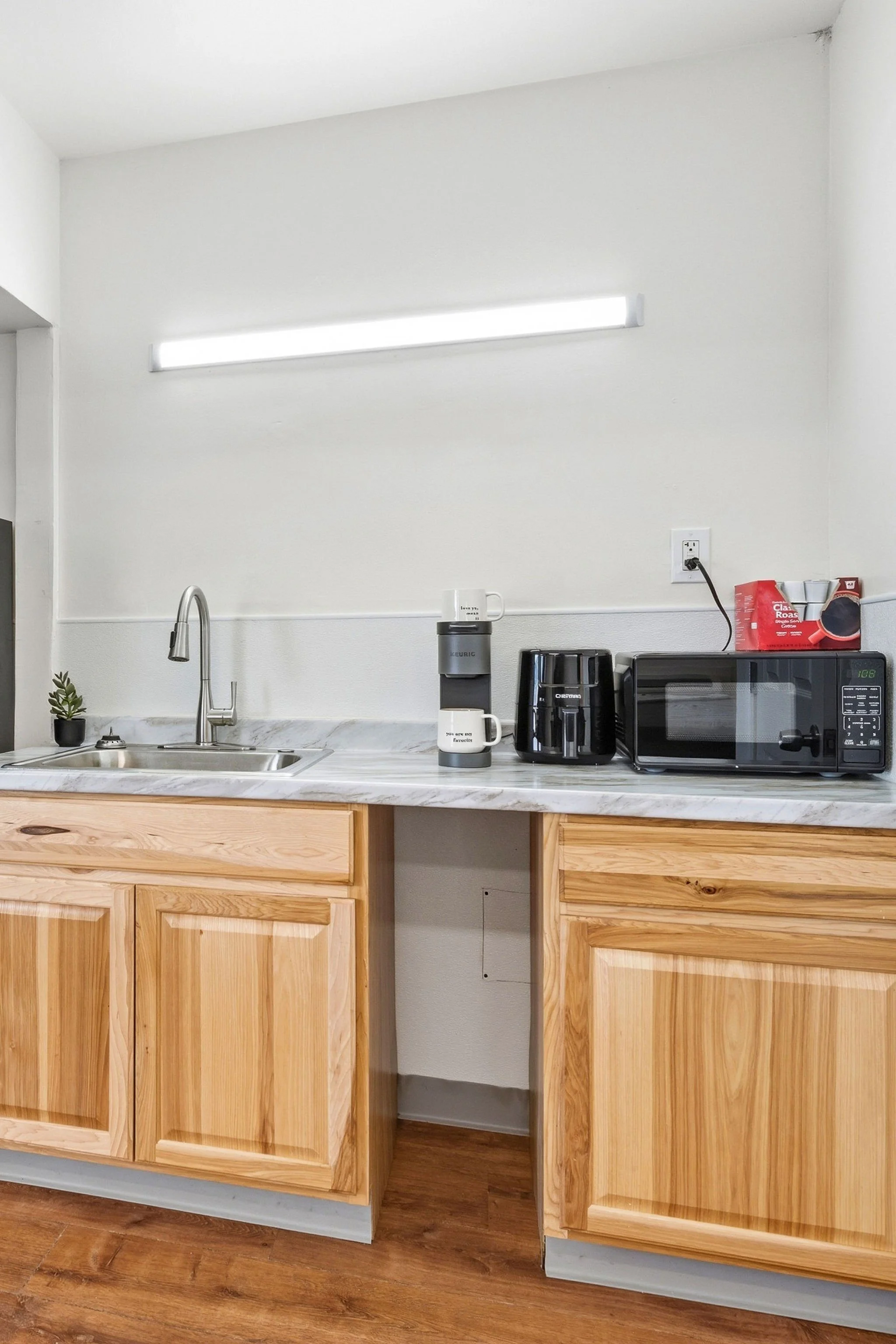 Kitchen with light wood cabinetry, white marble countertop, stainless steel sink, small plant, coffee maker, microwave, coffee filters, and boxed coffee on the counter, with a blank wall and illuminated light fixture above.