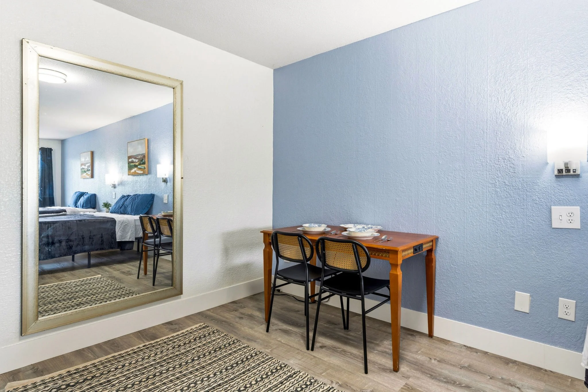 A mirror reflecting a bedroom with a bed, artwork, and curtains, beside a dining area with a wooden table, black chairs, and place settings with bowls and utensils, on a wood floor with a black-and-white woven rug, and a blue painted wall with a ther
