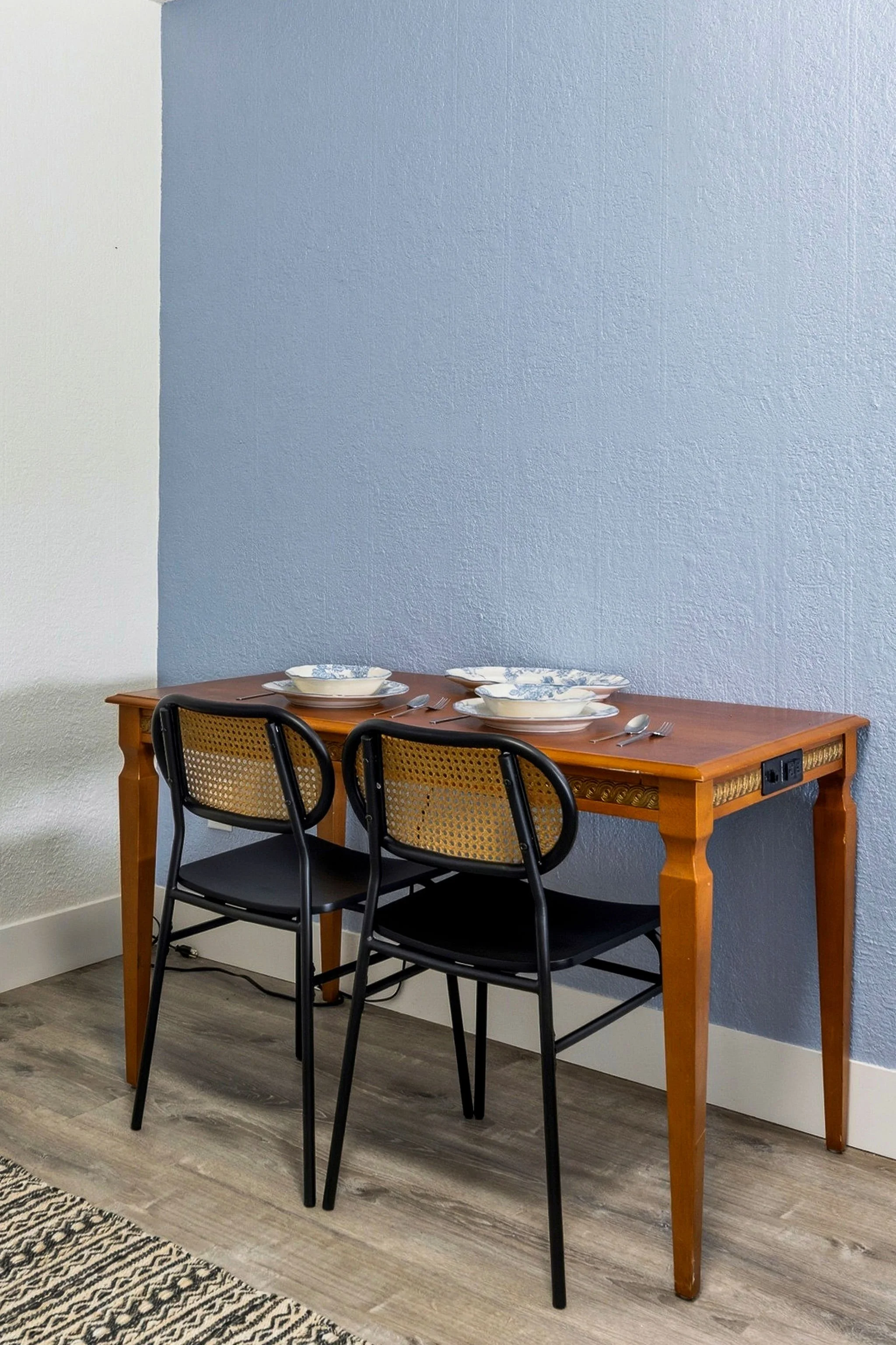 Wood dining table set for three with white and blue patterned plates, silverware, and napkins. Four black chairs with woven seats are positioned around the table. The background features a textured blue wall and a section of a plain white wall, with 