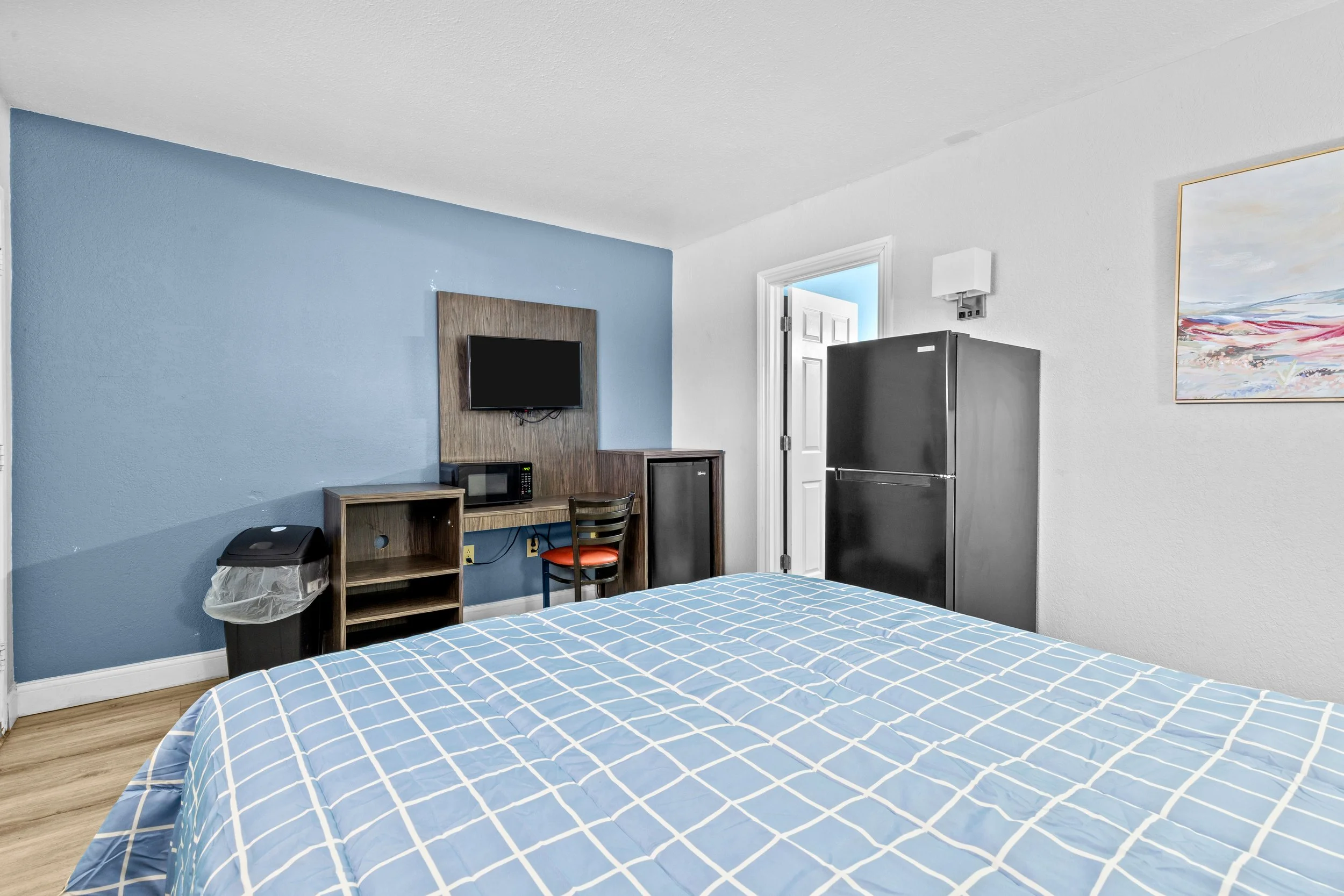 A bedroom with a blue accent wall, a bed with a blue and white checkered duvet, a small desk with a microwave and chair, a black refrigerator, a wall-mounted TV, a framed landscape painting, and a doorway leading to another room.