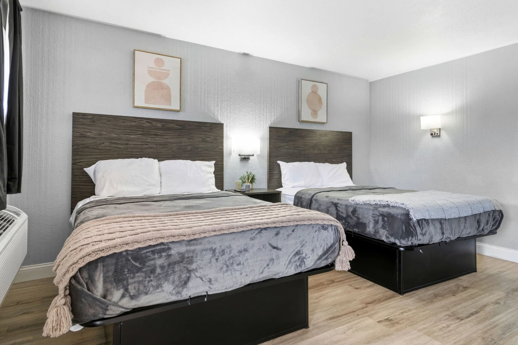 A hotel room with two beds, each with a wooden headboard, white pillows, and gray bedding. There is a side table between the beds with a small plant and a lamp. The walls are white, and there are two abstract framed artworks hanging above the beds. A