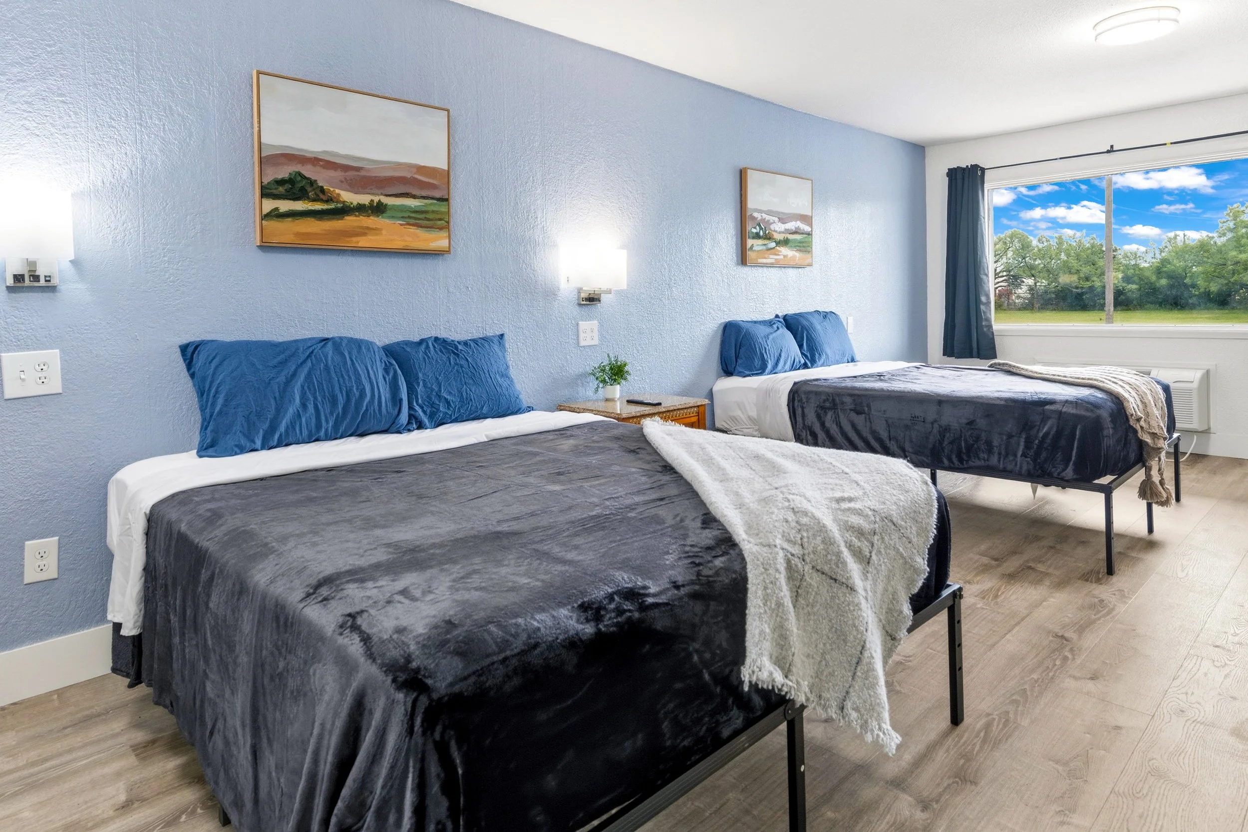 Room with two beds, each with blue pillows and dark gray blankets, bedside table with a small plant, artwork on the light blue wall, large window showing green trees and a blue sky.