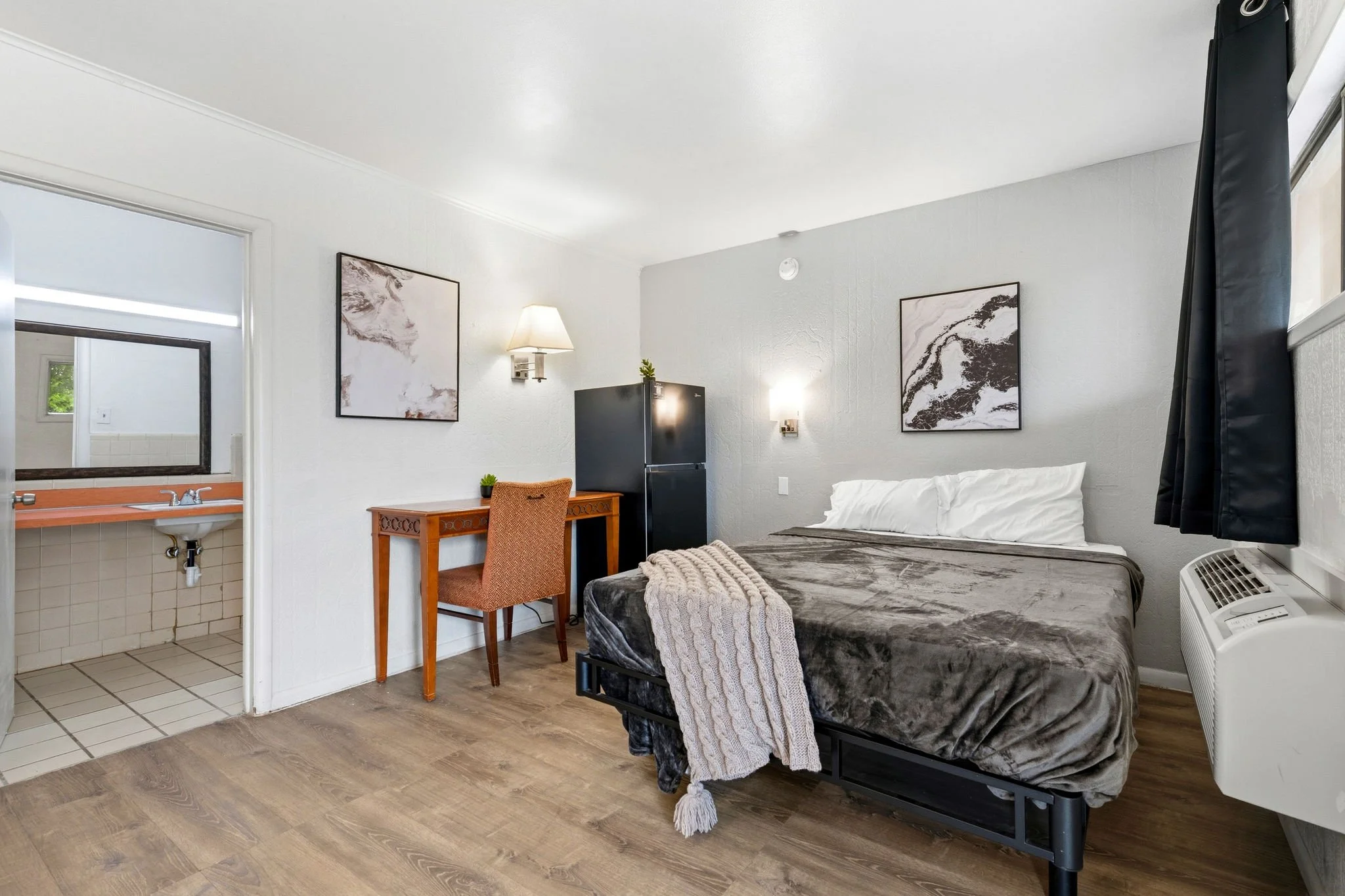 A compact hotel or motel room with a bed, artwork on the wall, a small fridge, a wooden desk with a patterned orange chair, a wall-mounted lamp, and an adjoining bathroom with a mirror and sink. The room has light-colored walls and wood flooring.