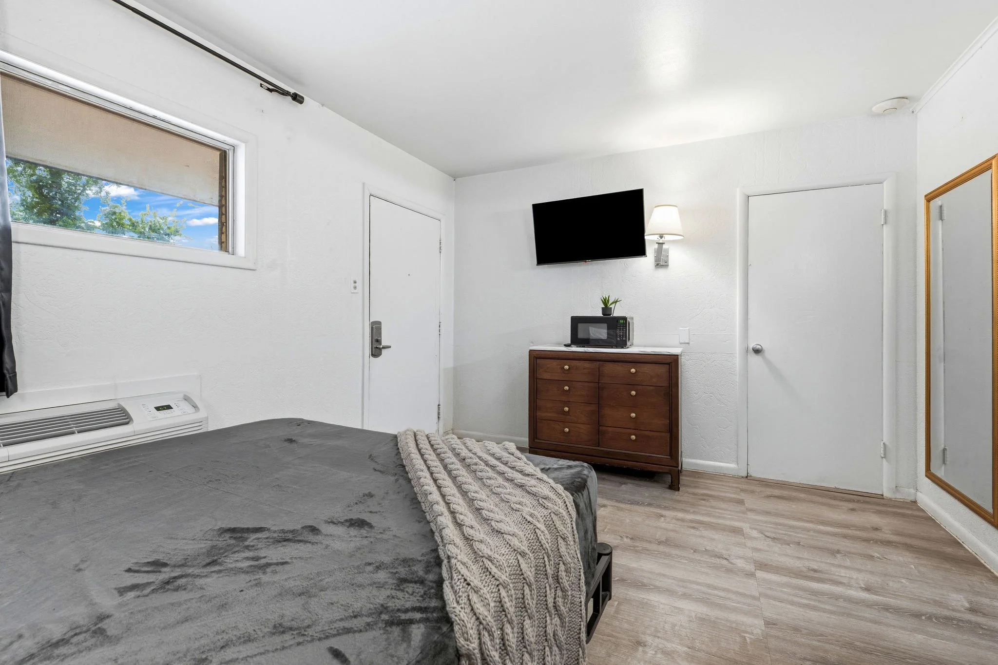 A bedroom with white walls and two doors, a window showing green trees and blue sky, a wall-mounted TV, a lamp, a microwave on a wooden dresser, and part of a bed with a gray blanket and a knitted throw blanket.