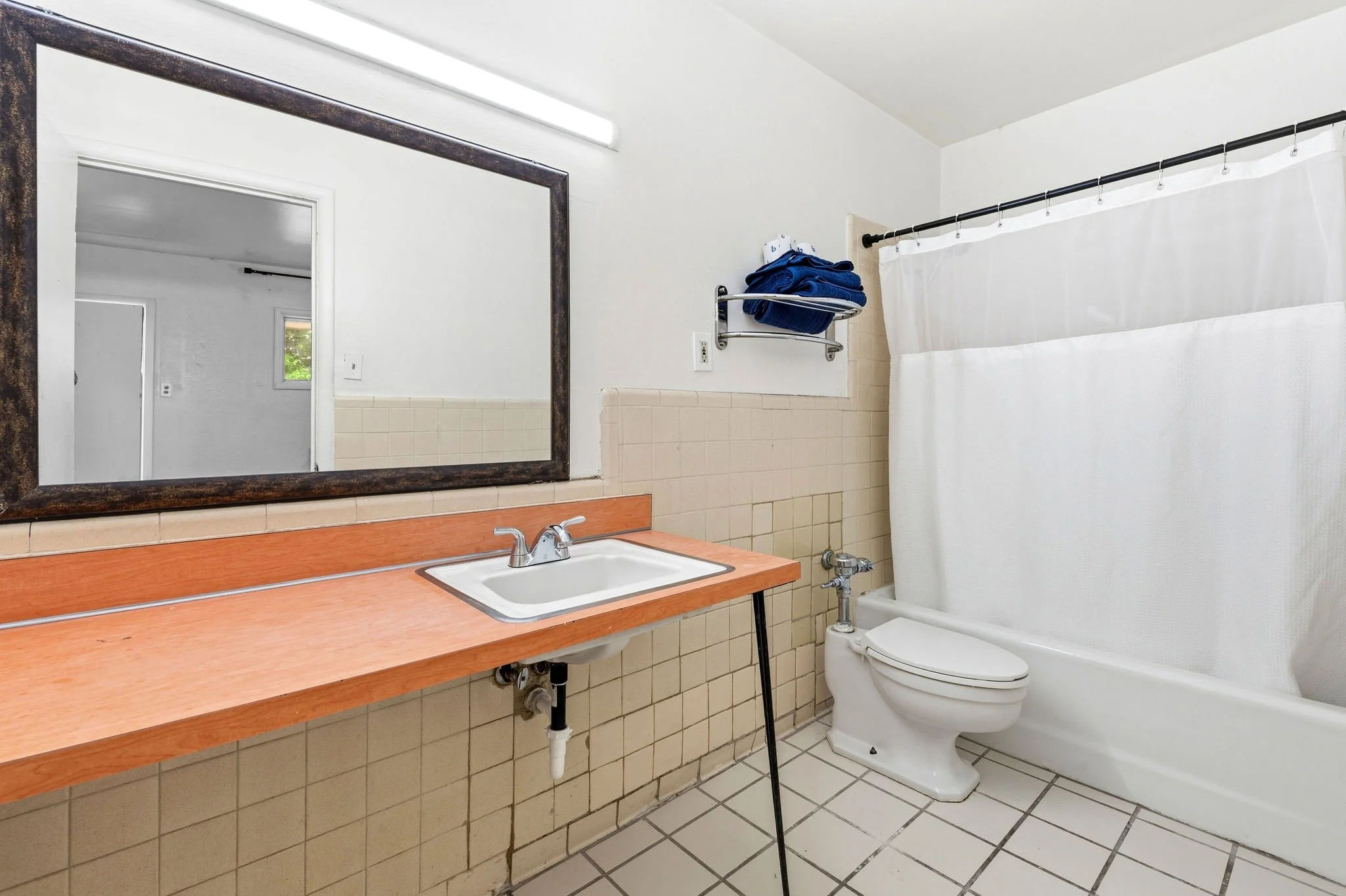A bathroom with a mirror, sink, towel rack with blue towels, toilet, shower with white curtain, and beige tiled walls and floor.