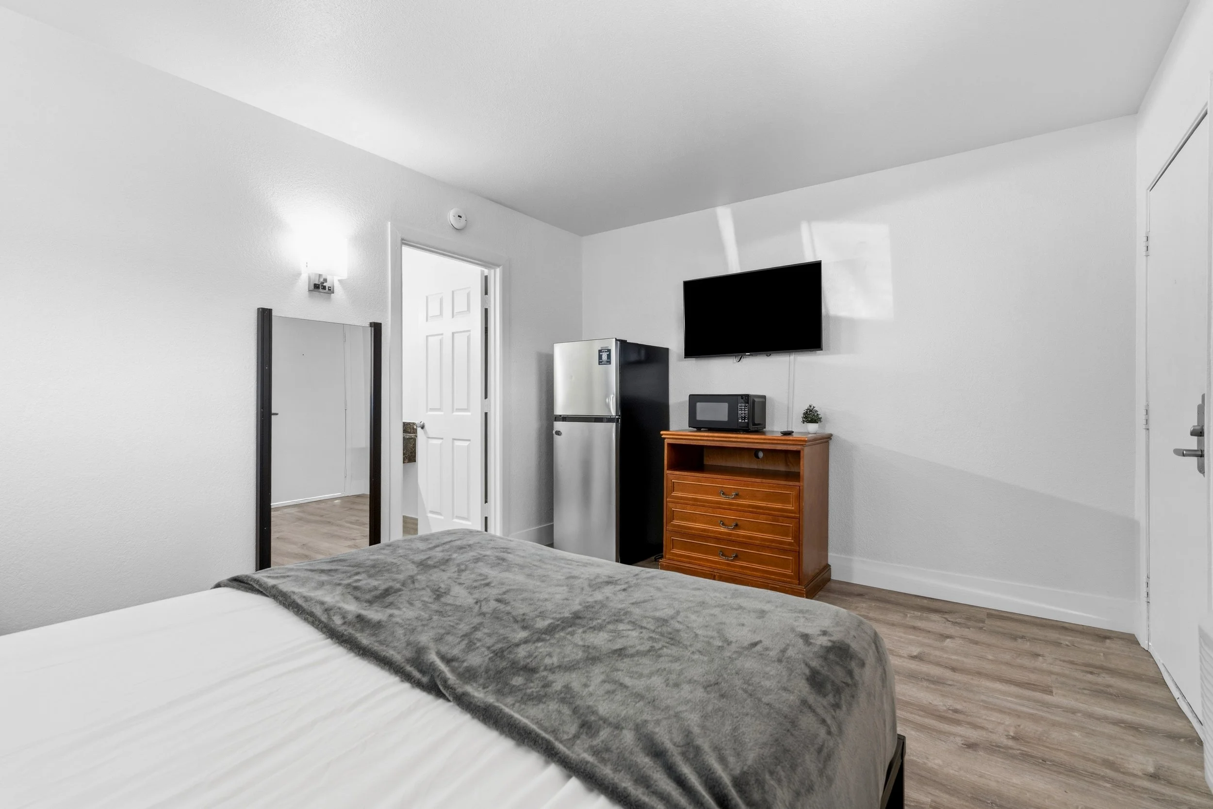 View of a bedroom with white walls and wood flooring, showing part of a bed with gray blanket, a small wooden dresser with drawers, a black mini fridge, a microwave, a wall-mounted TV, a door leading to another room, and a closet door.