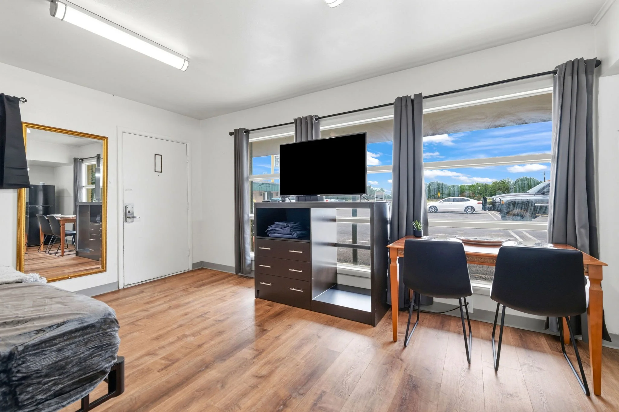 Interior of a small apartment or studio with wooden flooring, a large window with gray curtains, a TV on a stand, a wooden table, and chairs. Outside the window, a parking lot with cars and a bright blue sky with clouds are visible.