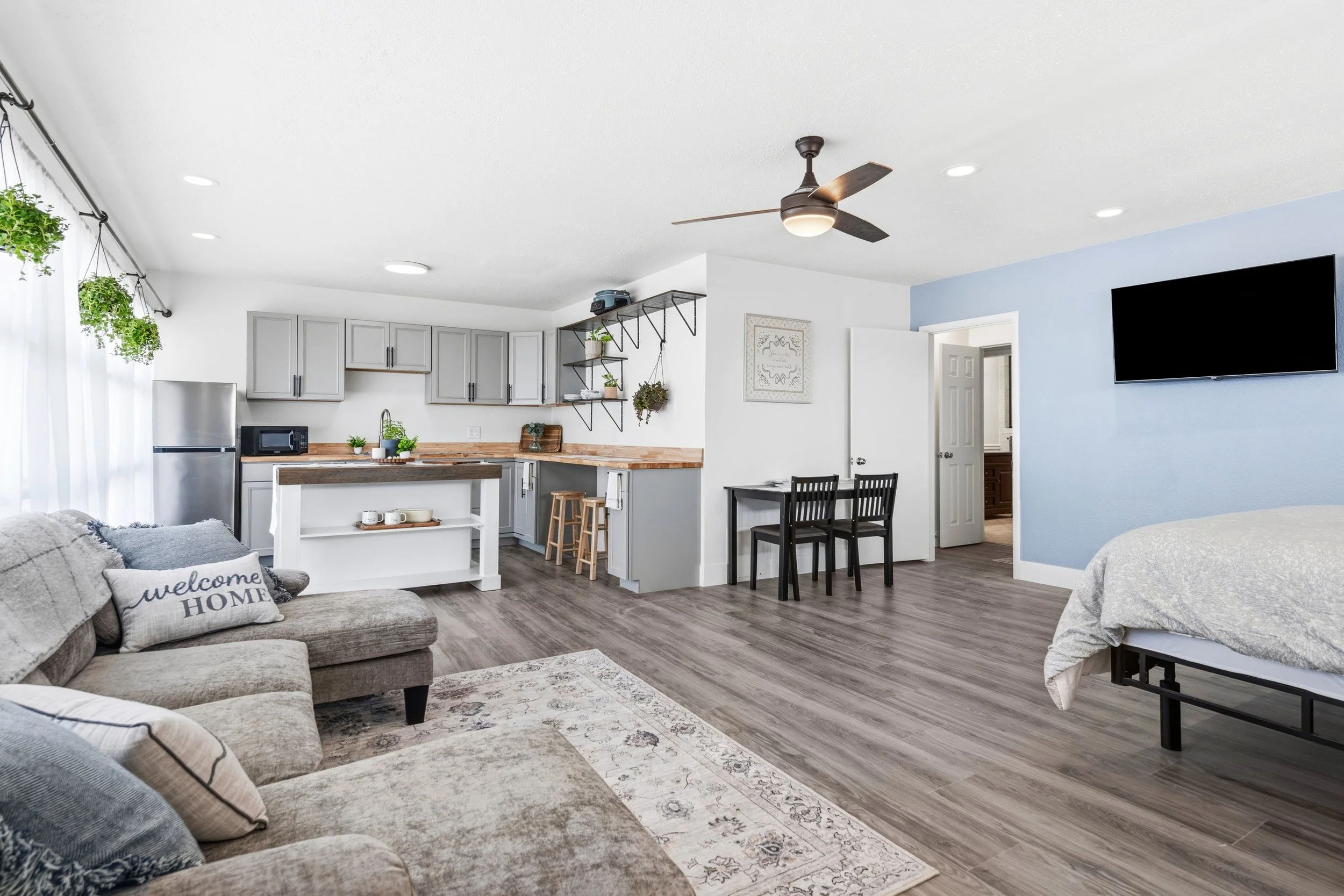 Living room with gray sofa, decorative pillows, light wood flooring, open kitchen with gray cabinets, black bar stools, mounted TV on blue accent wall, ceiling fan, and hanging plants.
