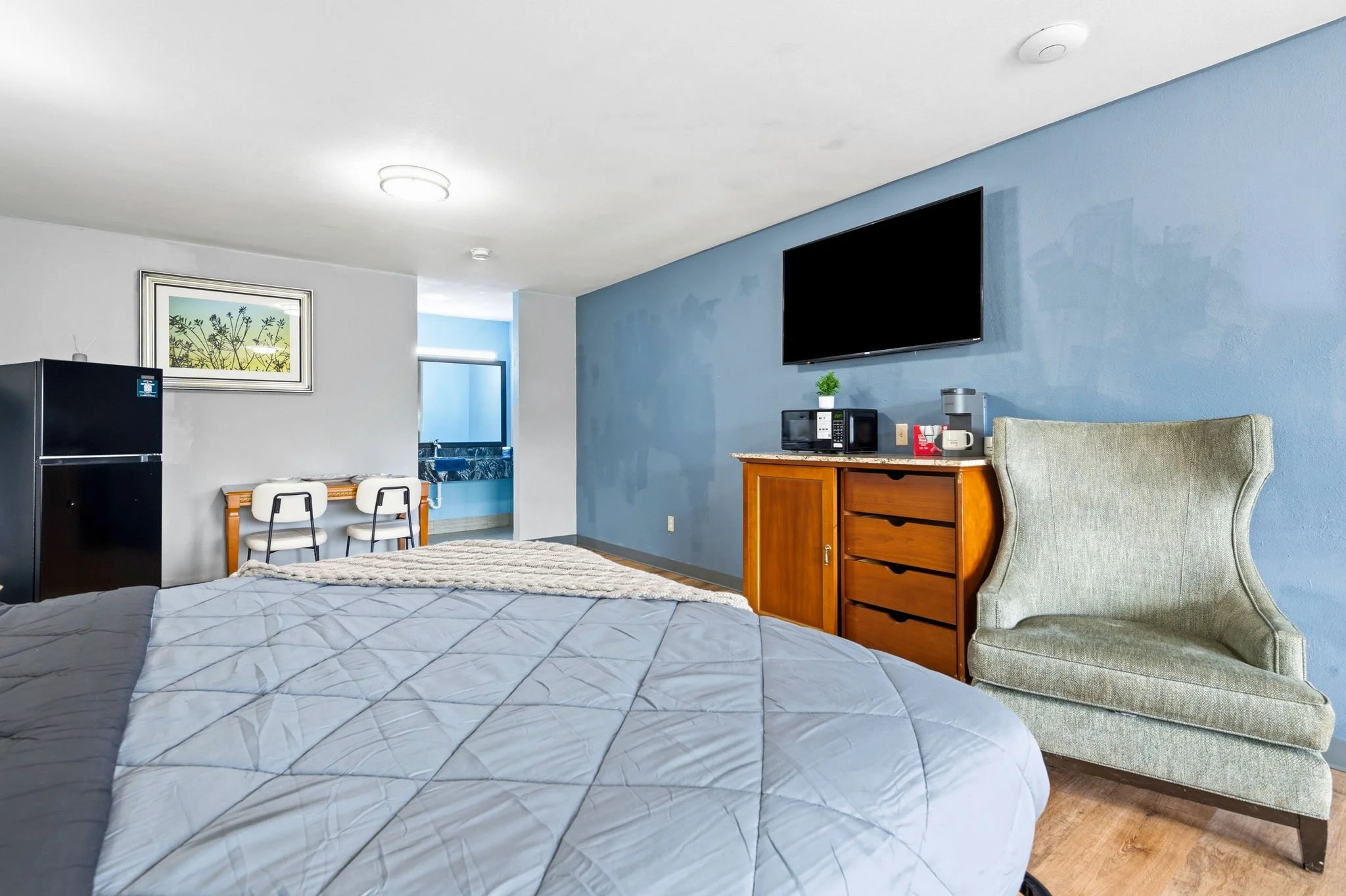 Interior view of a hotel room with a bed in the foreground, wooden furniture, a flat-screen TV mounted on a blue wall, and a small kitchenette area with a refrigerator and microwave.