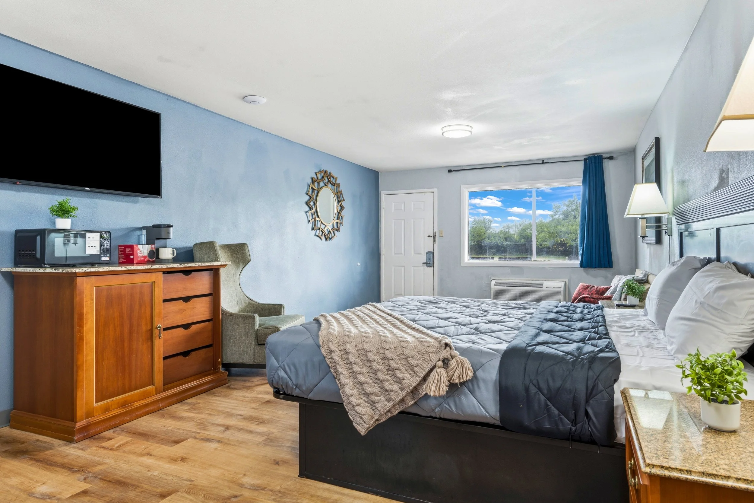 A cozy hotel room with a bed, window, and various furnishings. The room features a large bed with a quilted gray bedspread and decorative pillows. There is a small bedside table with a potted plant and a lamp. On the opposite wall, a flat-screen TV is mounted above a wooden dresser with a microwave, coffee maker, and a mug. A green armchair is next to the dresser. The room has light blue painted walls, a white ceiling with a ceiling light, and wood flooring. Outside the window, a clear blue sky with some clouds and greenery are visible.