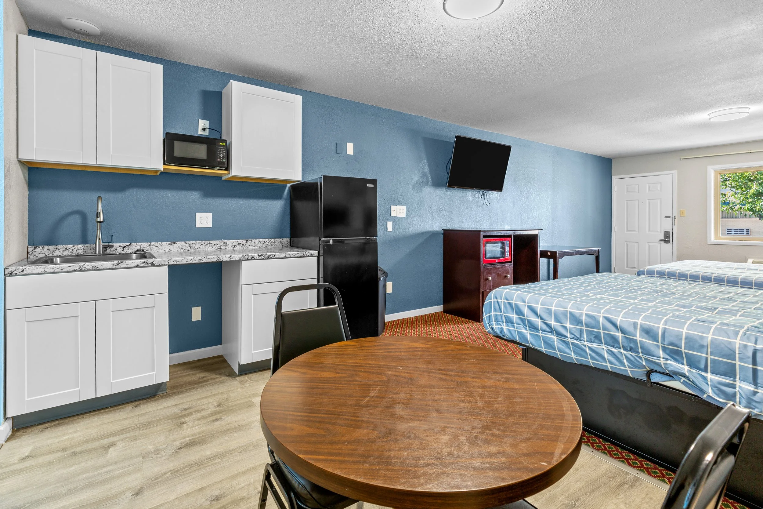 An interior of a cozy studio apartment with a kitchenette, a flat-screen TV mounted on the blue wall, a small black refrigerator, a microwave, a sink, two beds with checkered blue and white bedsheets, a round wooden dining table with four black chairs, and a window showing trees outside.