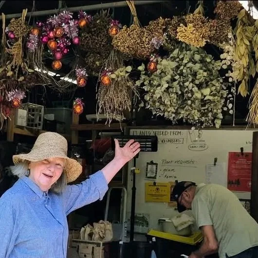 A woman wearing a large straw hat smiling and gesturing in a garden store, with hanging flowers and dried plants above her, while a man works at a counter in the background.