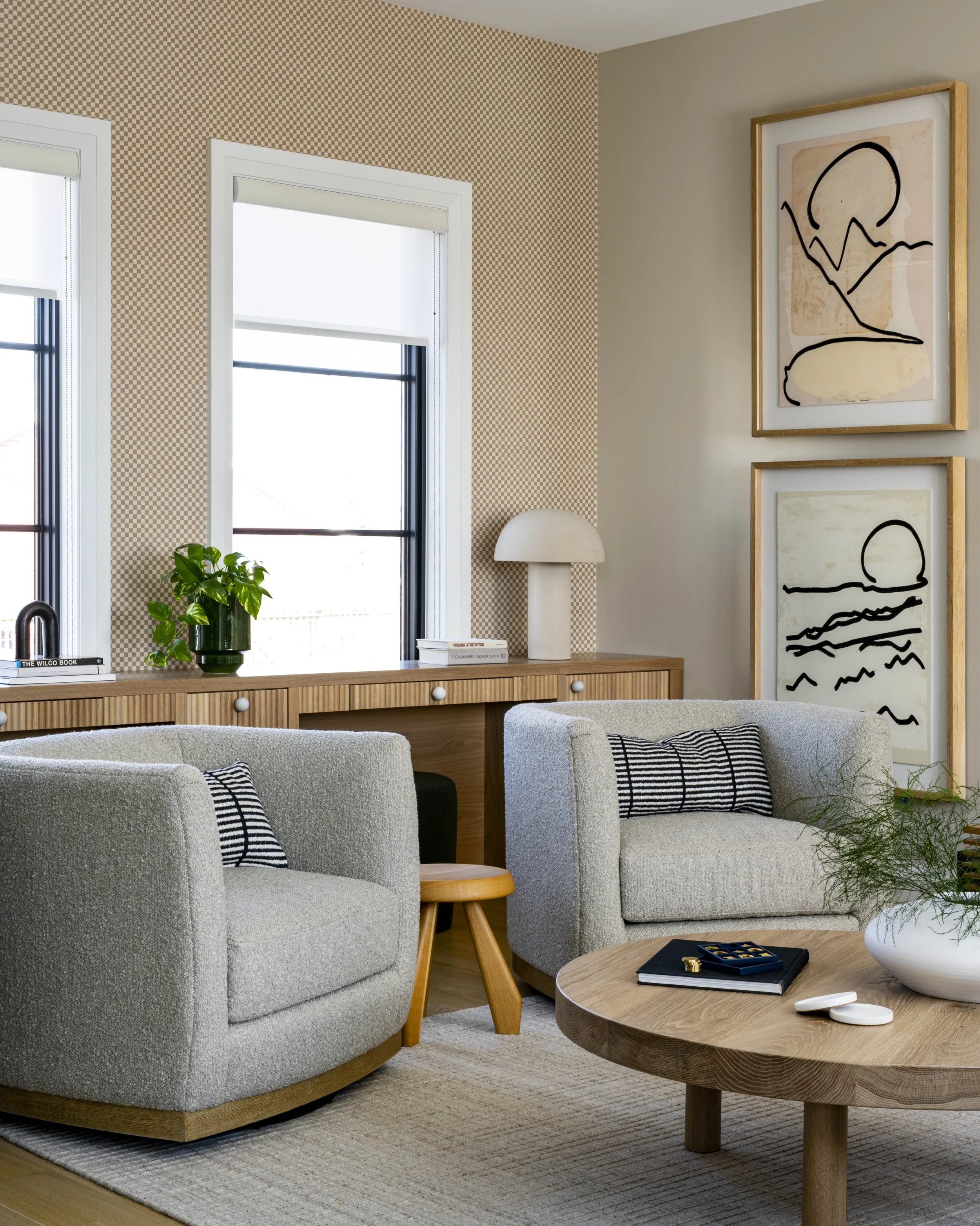 Living room with beige armchairs, a round wooden coffee table, framed abstract art on the wall, two windows with white blinds, and decorative items including a plant and books.