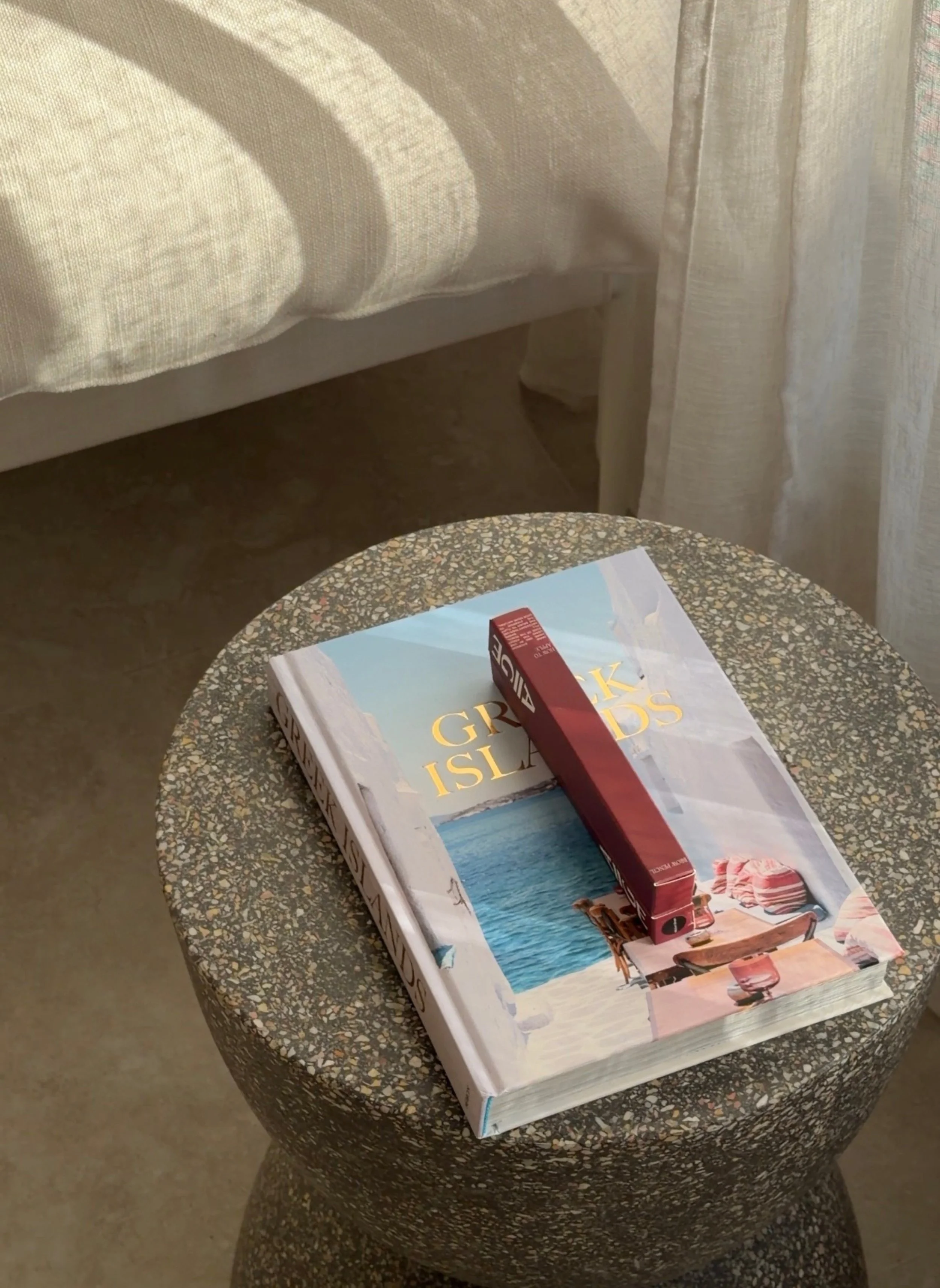 A small round grey side table with a 'Greek Island' coffee table book on top and two Aiige eyebrow pencils in their red boxes displayed on top.