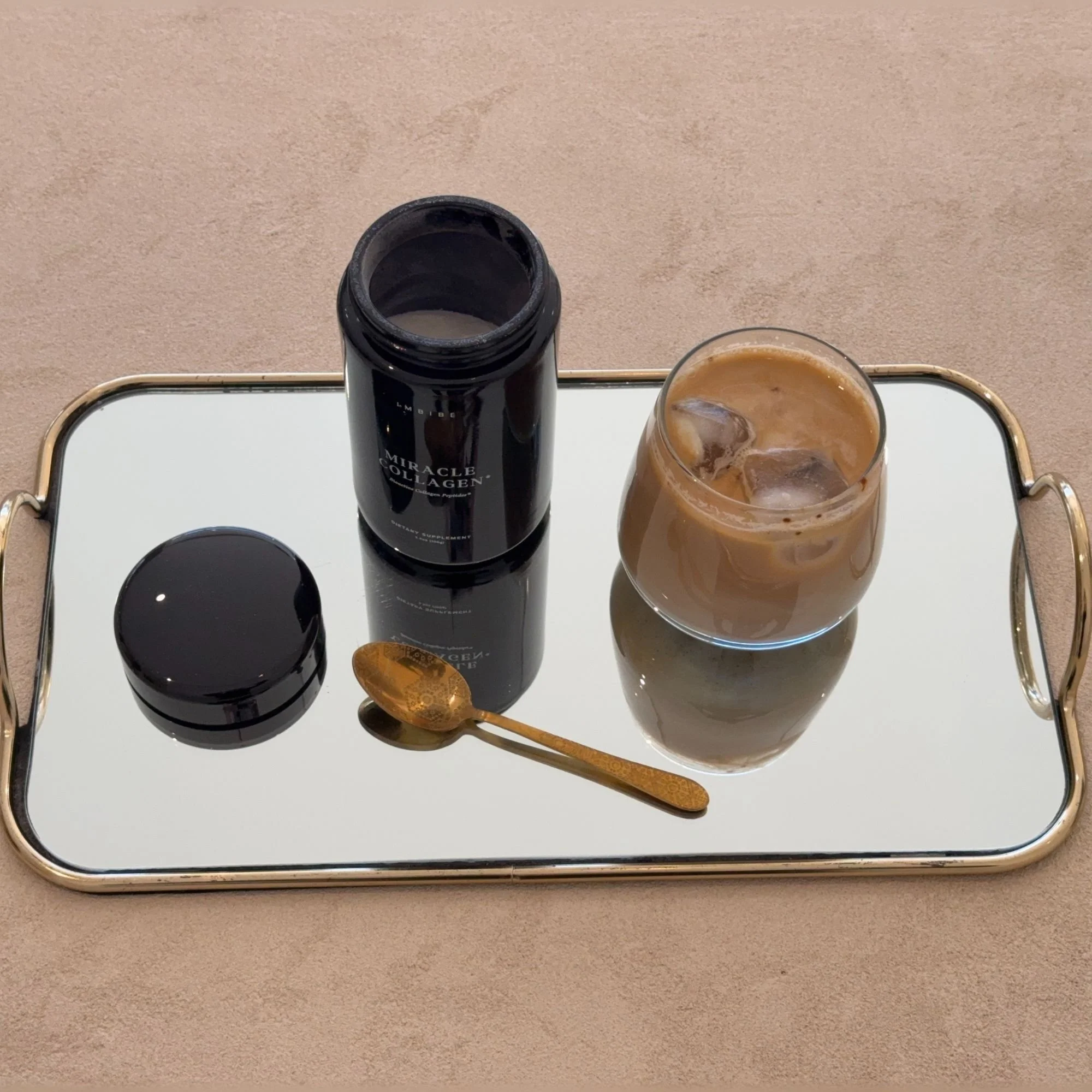 the 100 gram glass jar of IMBIBE's miracle collagen with an iced coffee next to it to stir in and enjoy. items are placed on a mirror tray.