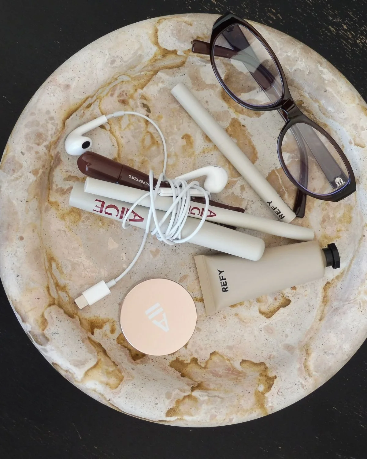 A marble dish holding an array of beauty products including AIIGE brow hold, brow sculpt and brow precise pencil.