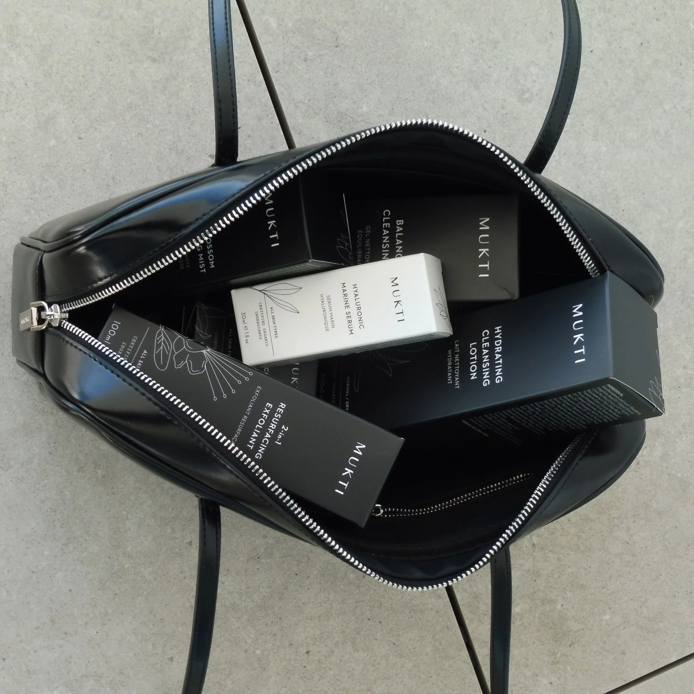 A black nakedvice handbag unzipped with Mukti Organics moisturiser and serum range inside stil in there black and white branded cardboard packaging.