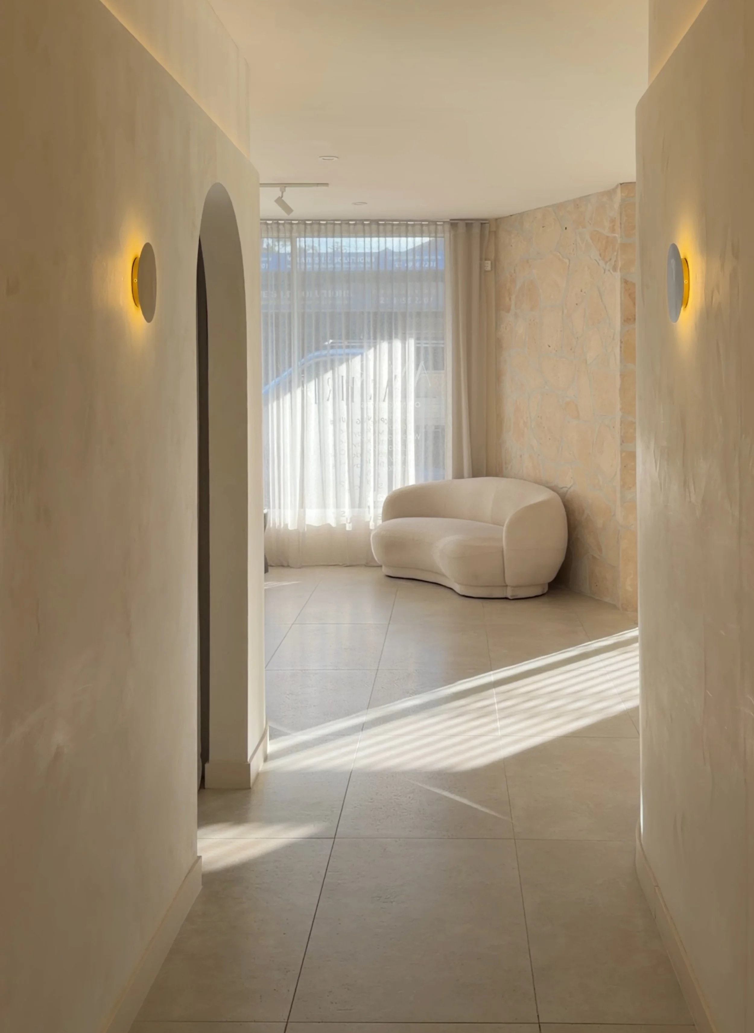 A naturally light filled space with travertine tiles and washed walls looking down the hall at a boucle lounge