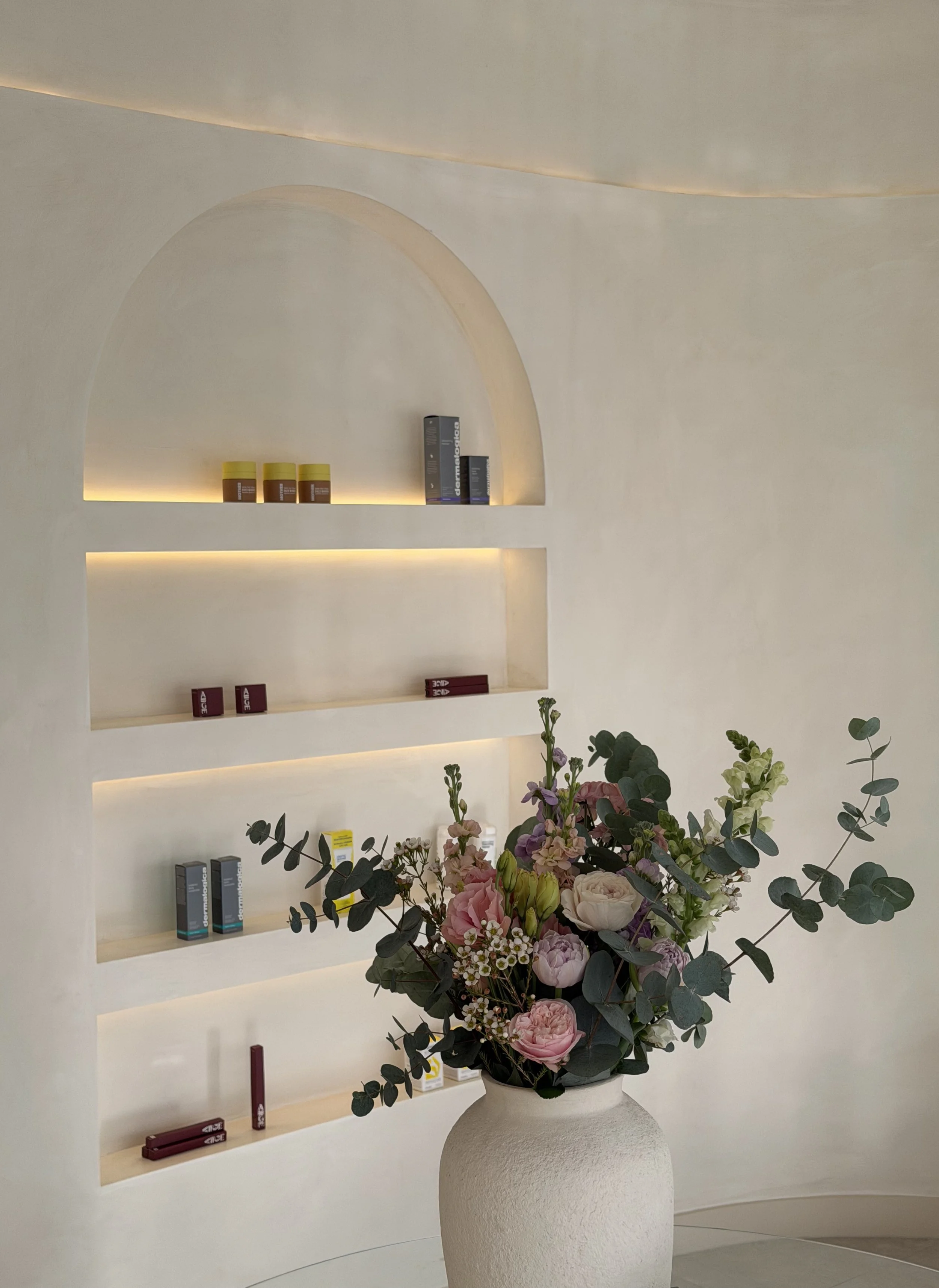 A curved product display wall in the background and a white vase with a fresh bunch of light pink flowers and eucalyptus leaves in amiris waiting room.