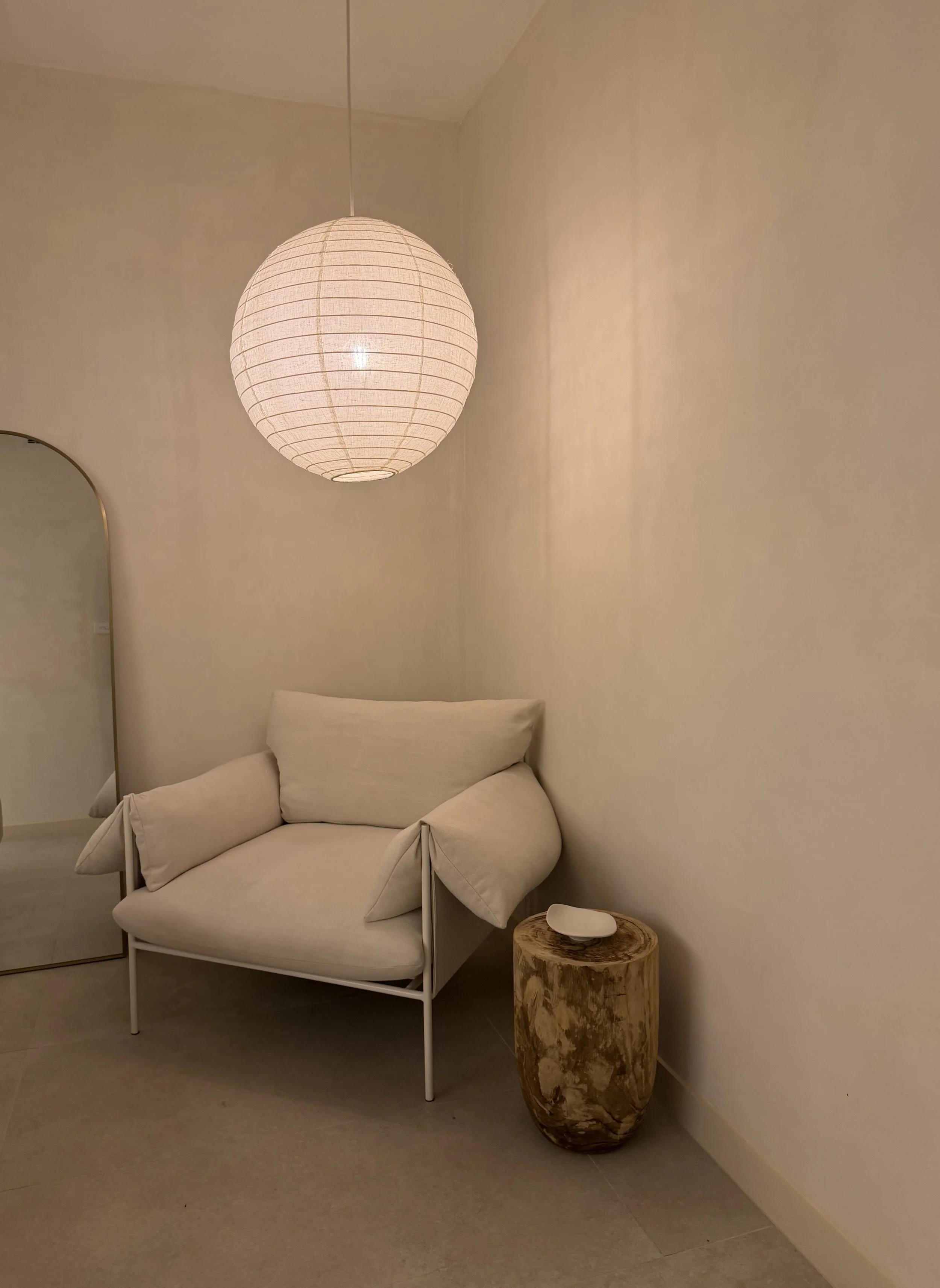 A linen lamp hanging from above with a large arm chair in the corner, wooden side table to enjoy
