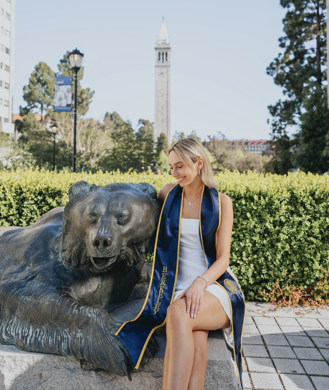 A woman in a white dress with a blue and gold graduation stole sits next to a bear statue in a park, with the bell tower of the University of California, Berkeley, in the background.