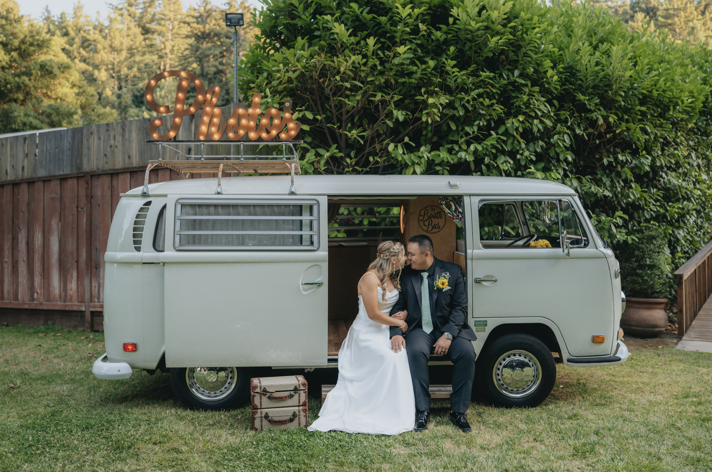 A bride and groom sitting inside a vintage white van with sliding door open, surrounded by green trees and grass, sharing a kiss. The van has a illuminated sign on top that says 'Photo!', and a suitcase is on the ground beside them.