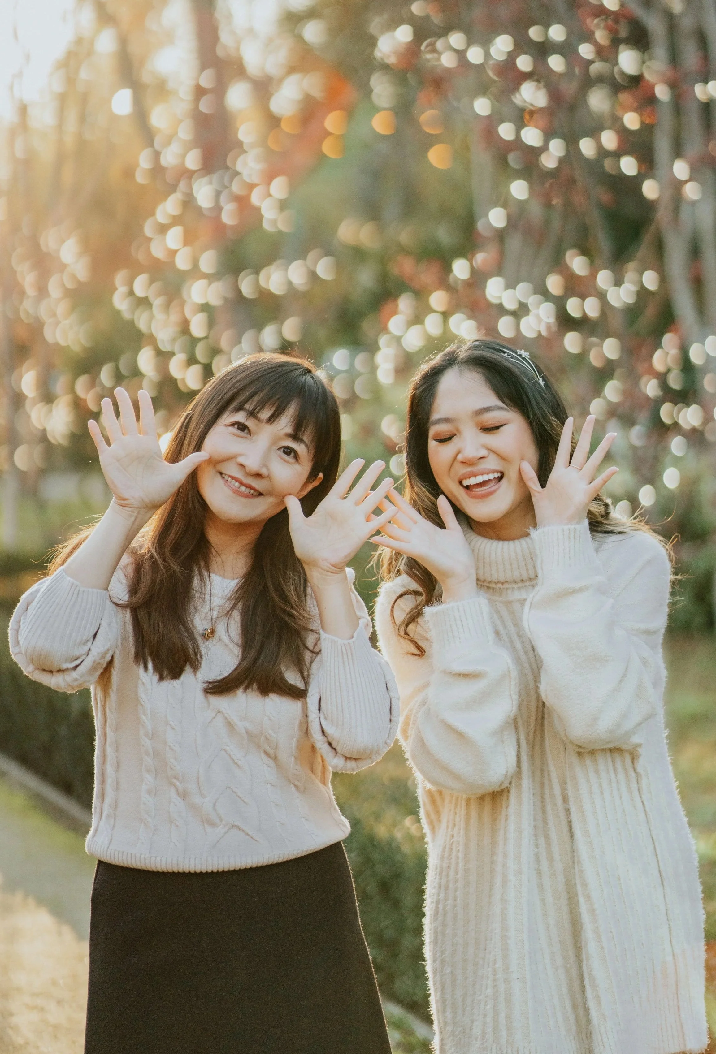 Two women smiling and waving at the camera in a park during sunset, with bokeh light effects in the background.