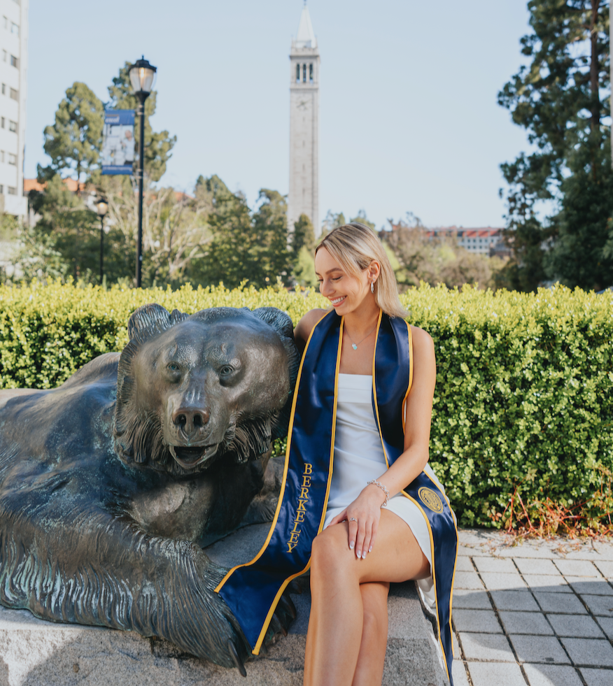 A woman in a white dress with a blue graduation stole and sash that reads 'Berkley' sits next to a bear sculpture in a park, smiling, with a tall clock tower and trees in the background.