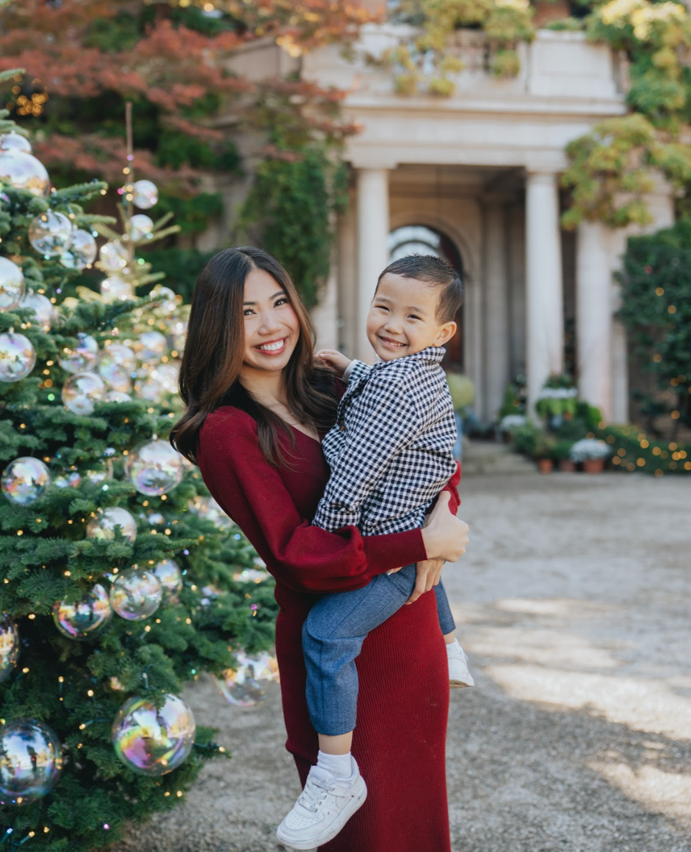A woman in a red dress holding a smiling young boy in front of a decorated Christmas tree outdoors during the holiday season.