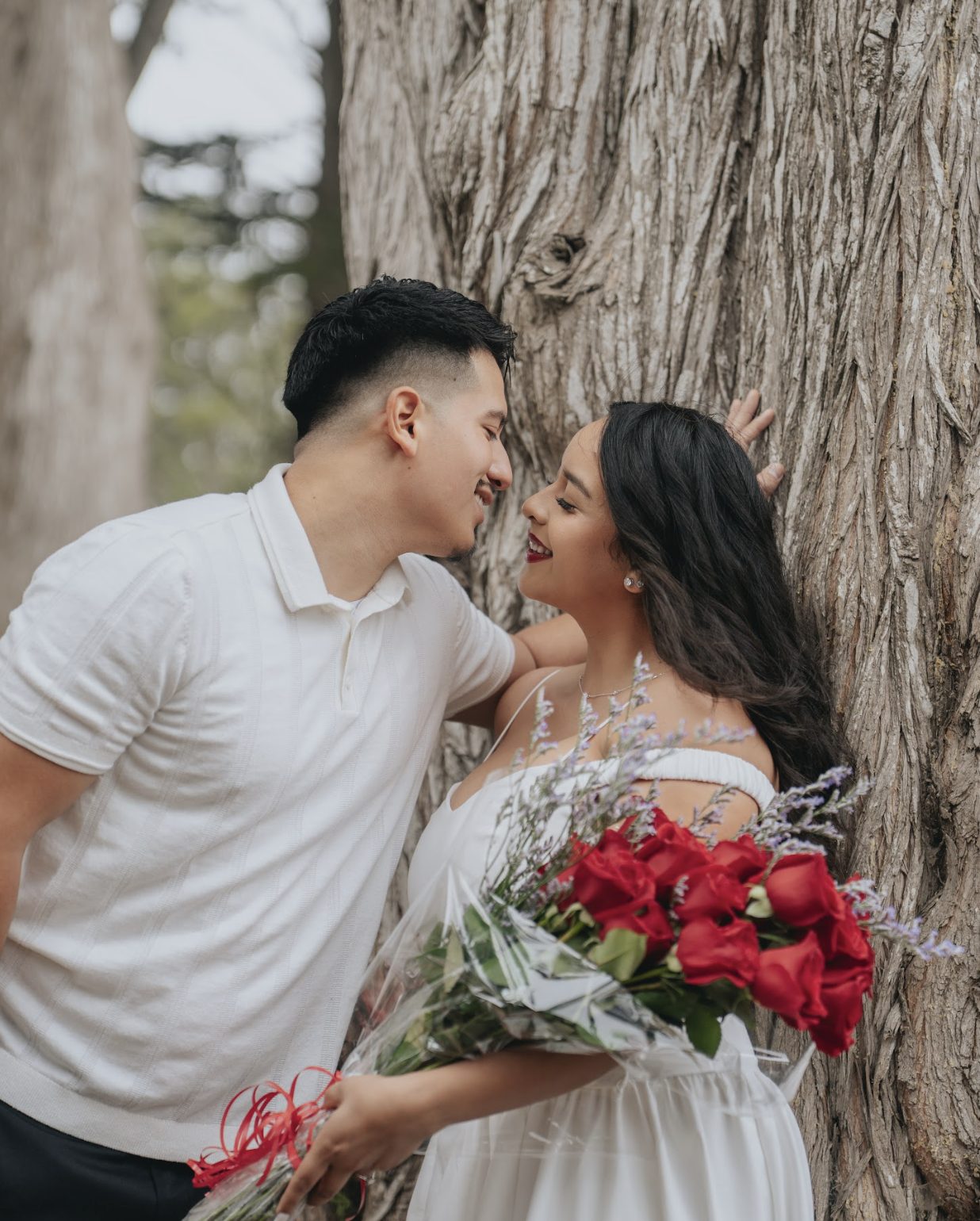 A romantic couple standing close to each other outdoors, with a large tree in the background. The woman is holding a bouquet of red roses and lavender, and they are smiling at each other.