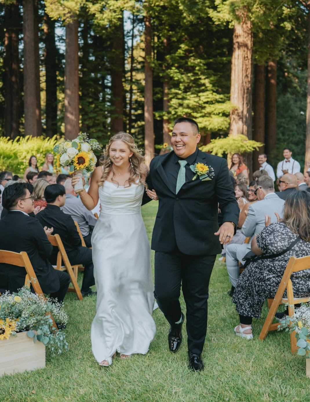 A bride and groom walking down the aisle at an outdoor wedding reception, smiling and holding hands, surrounded by seated guests in a forest setting with tall trees and green foliage.