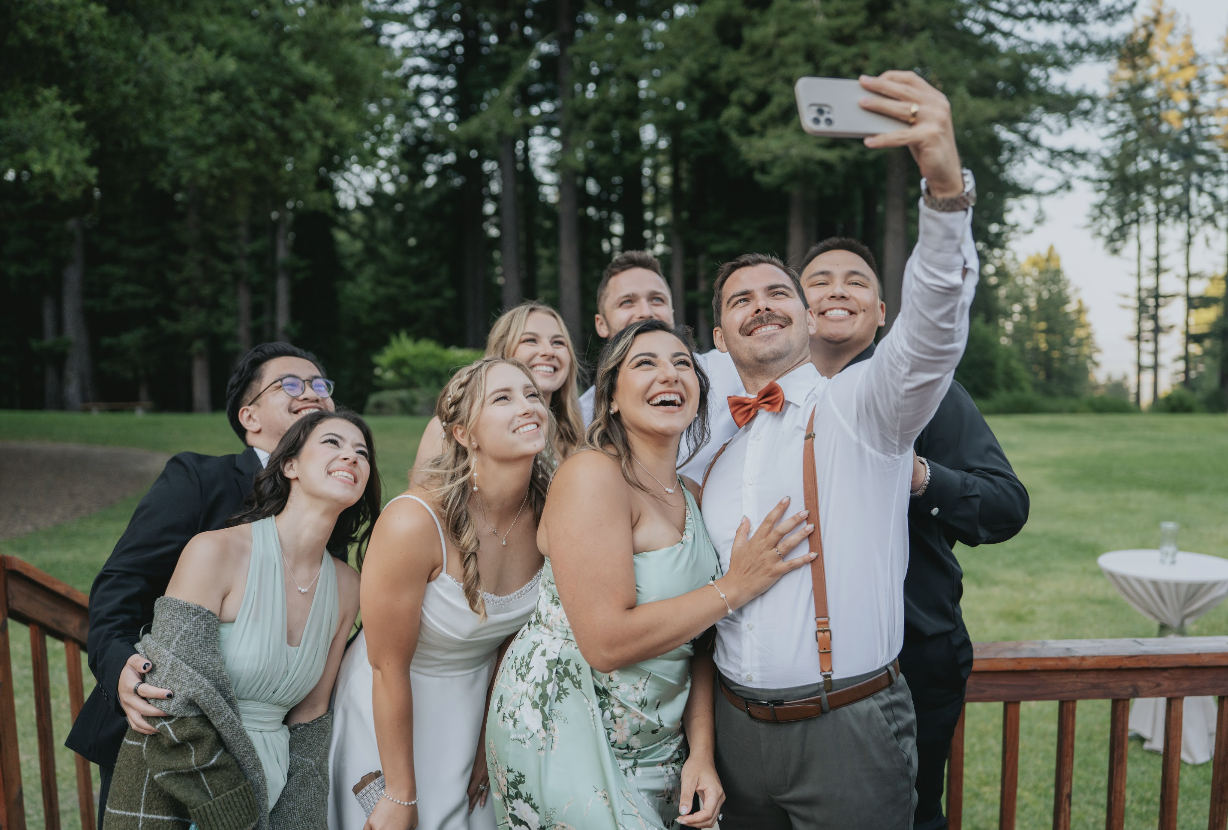 A group of eight people, dressed in formal attire, taking a selfie outdoors on a wooden deck with a green, wooded background. They are smiling and appear happy.