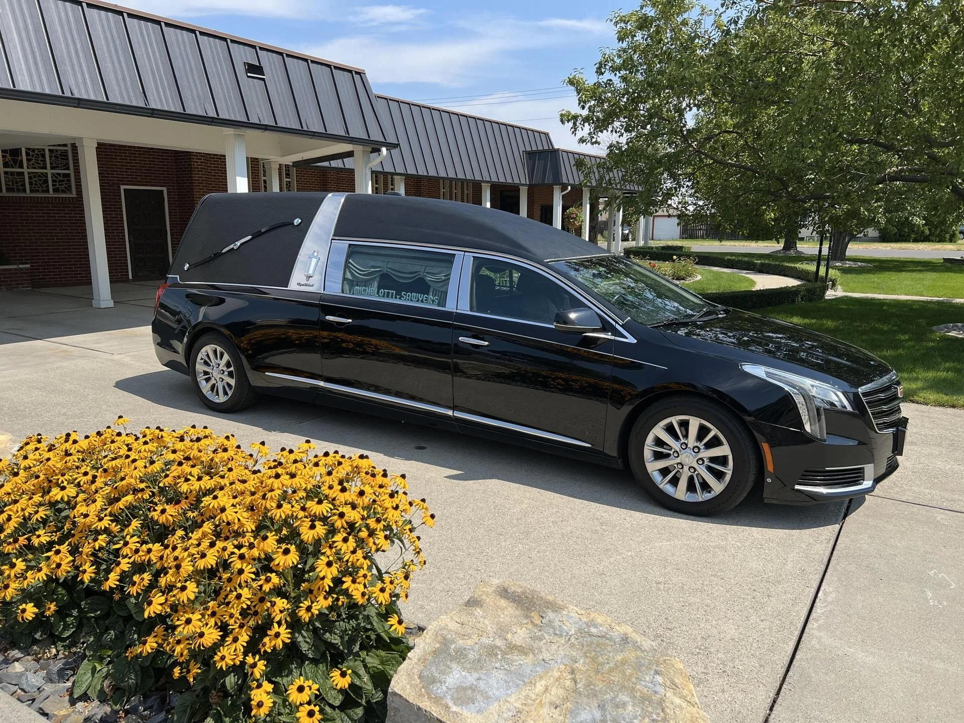 A hearse parked in front of a brick building with a metal roof, surrounded by green grass, trees, and yellow flowers.