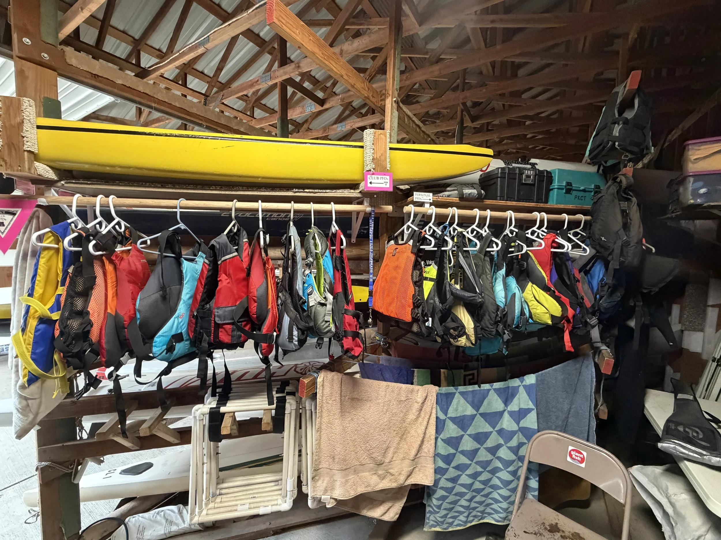 Garage storage area with a yellow kayak on a wooden shelf, backpacks hanging below, and various boxes and cloth items stored on shelves and in baskets.