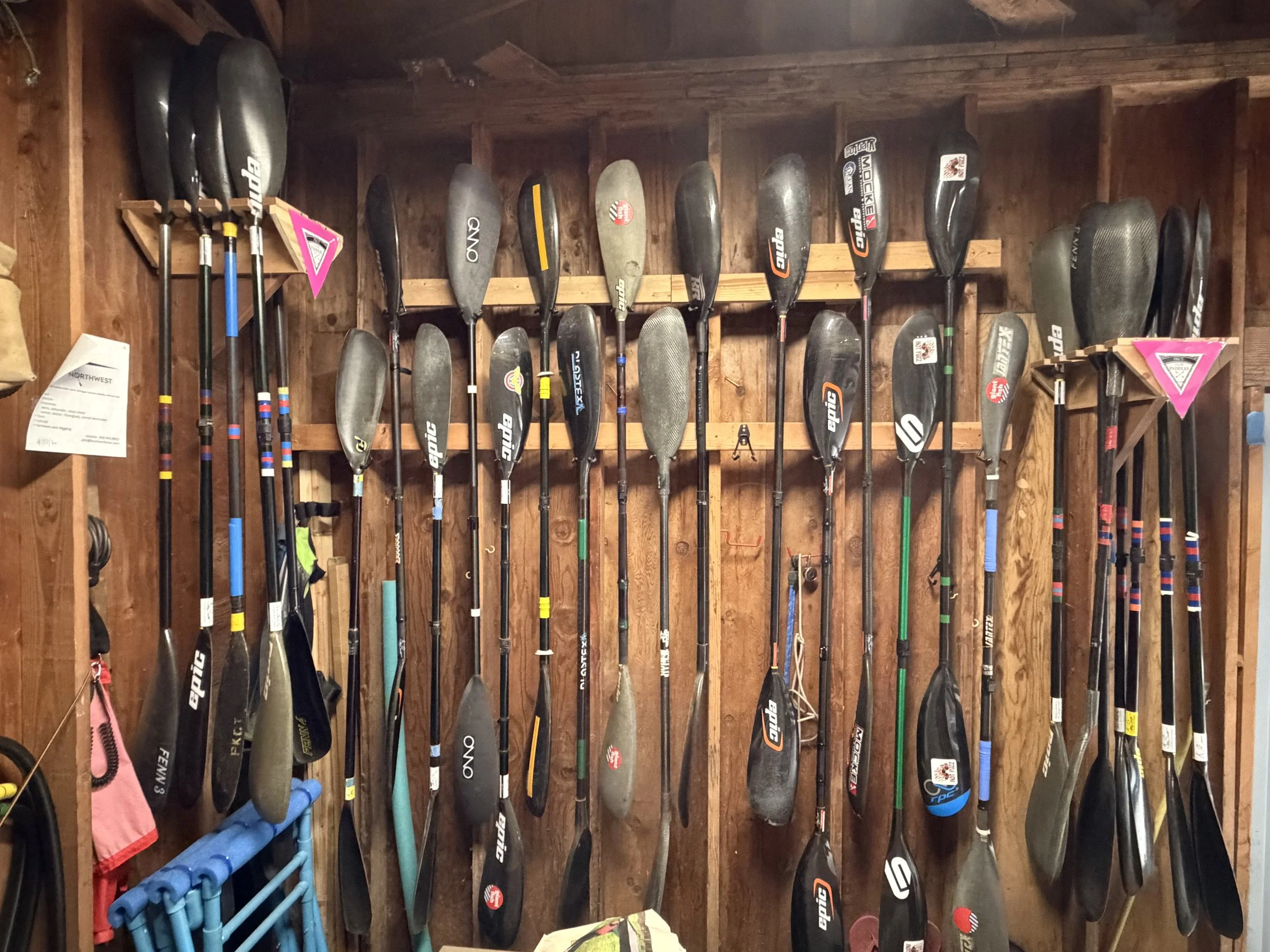 A wooden wall with multiple stand-up paddleboards hanging vertically, with a few paddle handles to the left.